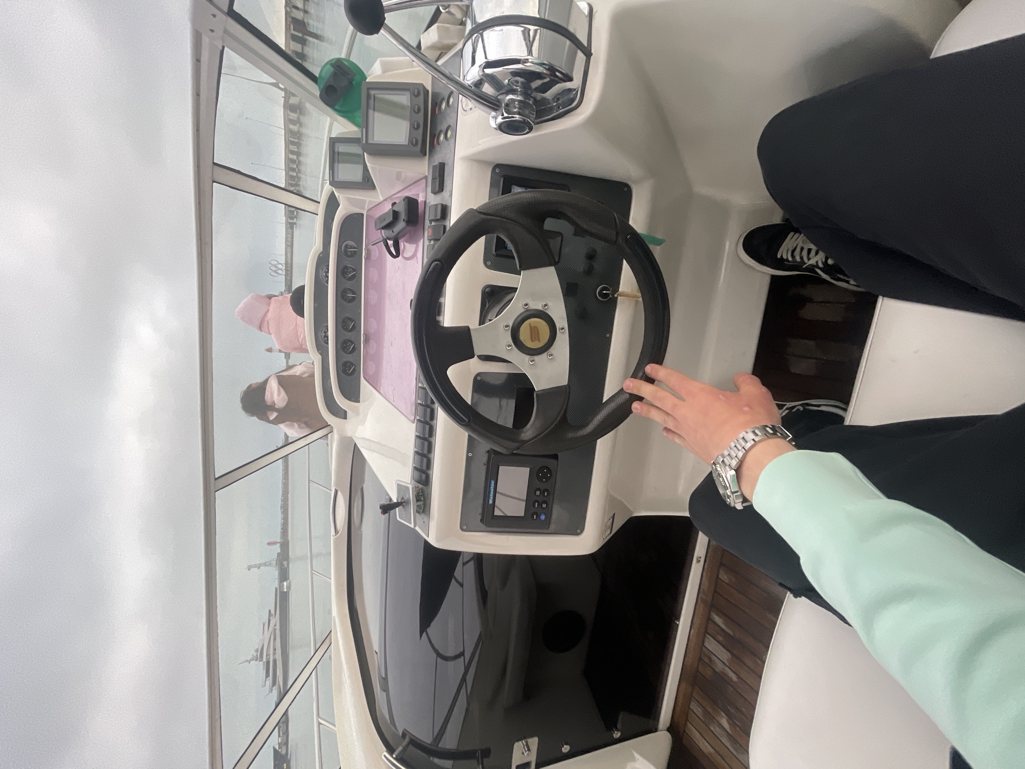 The interior of a boat cabin is shown. A person is at the helm, their hand on the steering wheel. To the left is a window showing a harbor with boats and buildings, potentially in Sochi, Russia, given the visible infrastructure and likely coastal location. The weather outside appears overcast. A second person is visible in the reflection in the window.