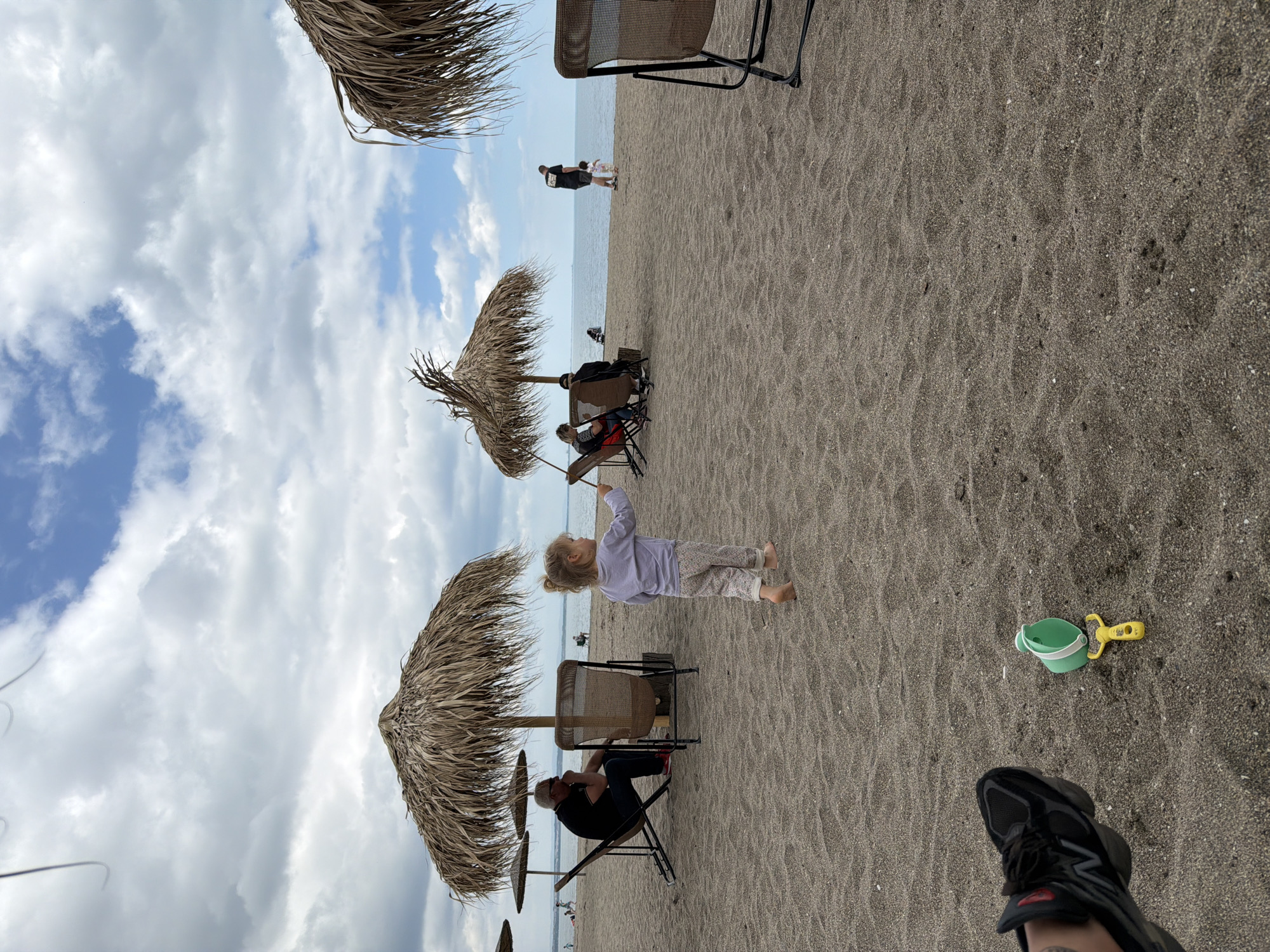 The image shows a beach scene in an unknown location. The sky is partly cloudy. Several thatch umbrellas are scattered along the beach. People are seen sitting in chairs under the umbrellas. A small child is standing on the sand, facing toward the horizon. In the foreground, a green bucket and yellow shovel are on the sand near a person's foot wearing a black sneaker. In the distance, two people are walking along the water's edge.