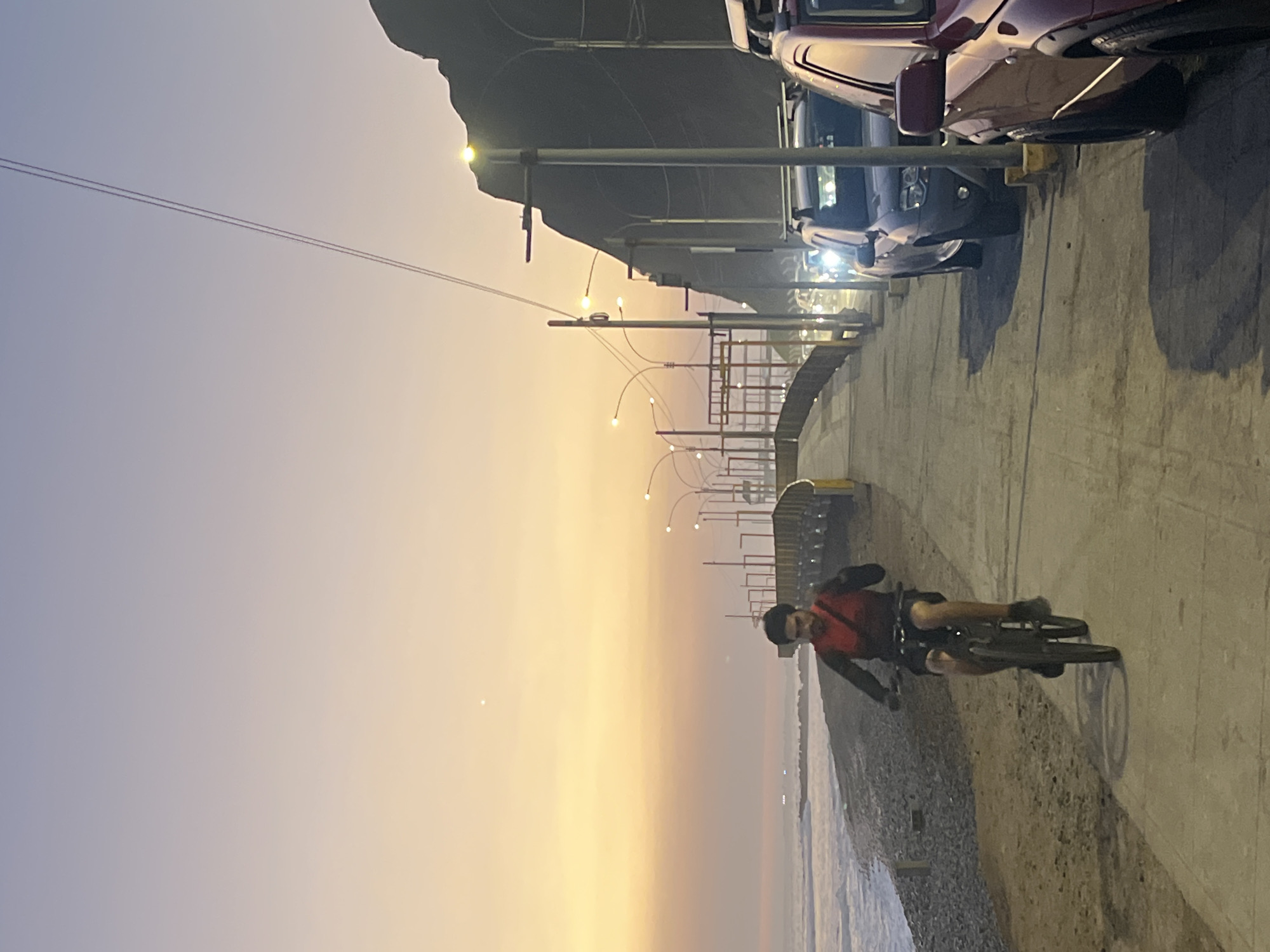 The image shows a man riding a bicycle along a paved walkway next to a beach in Miraflores, Peru. The man is wearing a red vest over a dark shirt and shorts, and is facing forward as he cycles. The walkway runs parallel to the Pacific Ocean, with waves visible on the left. In the background, there are streetlights lining the walkway and a steep, dark cliff face. Several cars are parked along the right side of the walkway. The sky displays a gradient of colors from pale yellow near the horizon to a soft purple above, indicative of either sunrise or sunset. A single bright point, possibly a planet or a star, is visible in the upper portion of the sky. The scene suggests an evening or early morning activity.