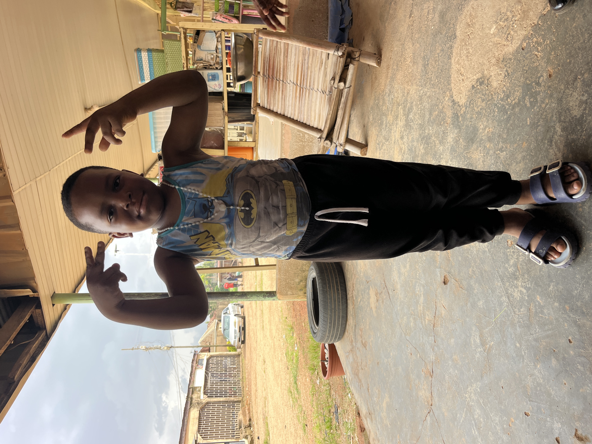 A young Black child, possibly male, stands centered in the frame, facing the camera directly. The child is wearing a blue and yellow sleeveless shirt with a Batman graphic and a white beaded necklace. Black pants are paired with dark blue sandals. The child's hands are raised, with fingers forming a gesture, and their expression is neutral.
The background suggests an outdoor, possibly rural or semi-urban setting. A covered porch area is visible, with a corrugated metal roof and wooden beams overhead. To the left, a dusty path leads to buildings with barred windows, and a white car is parked on the dirt. A used tire and a terracotta pot with a small plant sit on a concrete floor in the foreground. To the right, a rustic bamboo chair with woven slats is positioned, and shelves with various items are stacked behind it. The sky is overcast. The overall impression is a candid, everyday scene.