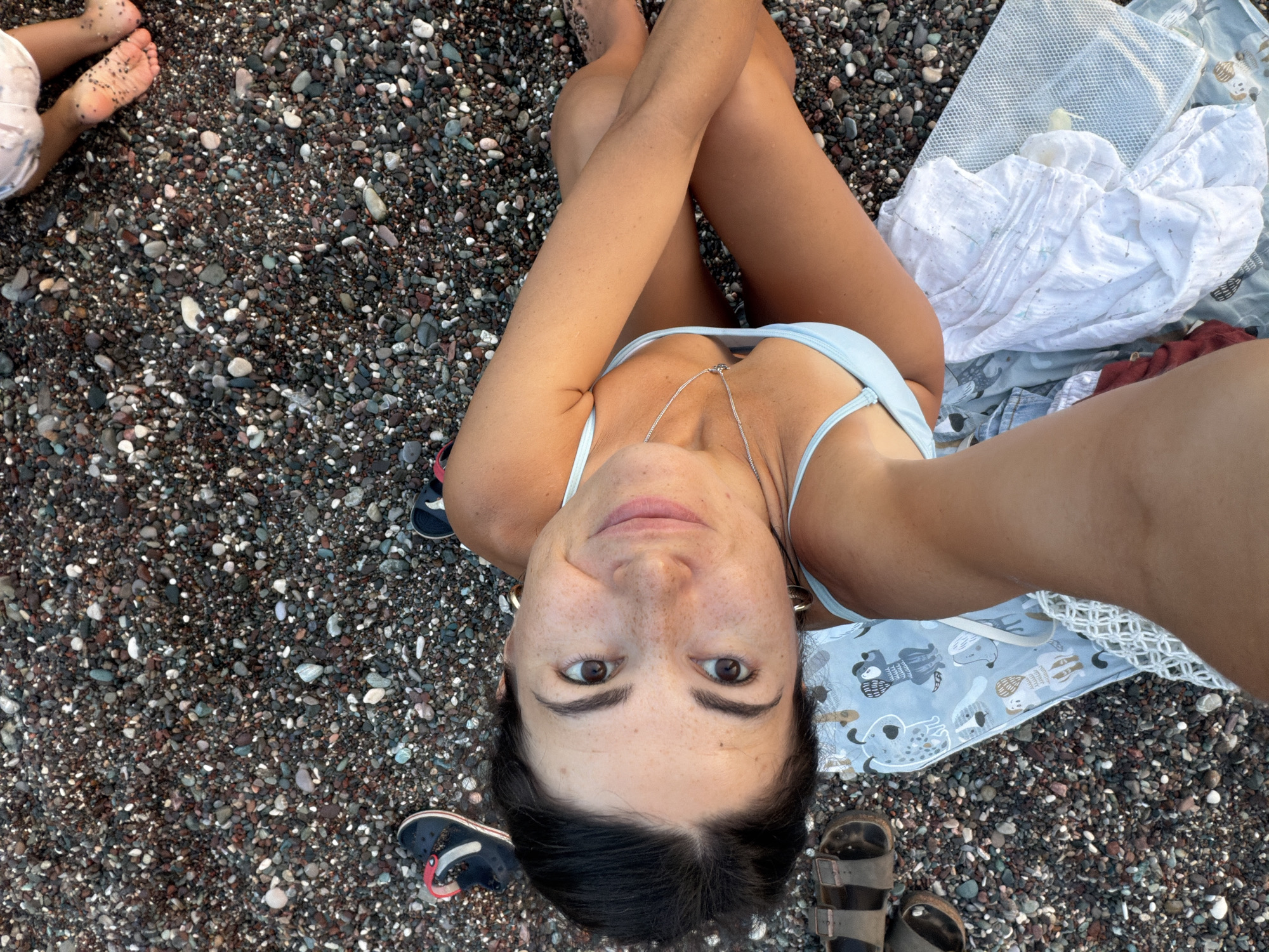 The image shows a woman sitting on a pebbled beach in Boreti, Montenegro. She is wearing a light blue bikini and has a necklace. Her legs are crossed, and she is looking up at the camera with a smile. There are also some children's feet visible in the upper right corner of the image, suggesting that the woman might be with her family. A patterned blanket is spread out on the beach next to her, and a white towel is also present. A pair of sandals is visible on the left side of the frame. The beach itself is covered in dark gray and brown pebbles of various sizes. The lighting suggests it is daytime.