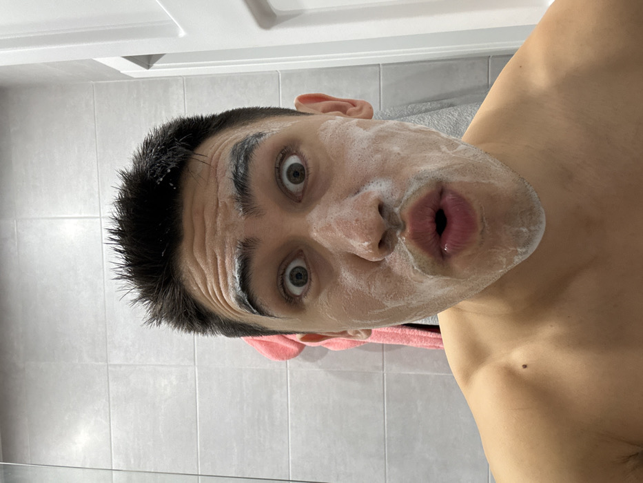 A shirtless man with black hair and fair skin gazes directly at the camera with wide, expressive eyes. Soap foam covers his face. His mouth is formed into an 'O' shape. He is inside of a bathroom. Behind him is the grey tiled shower. A partially visible pink towel can be seen in the background.