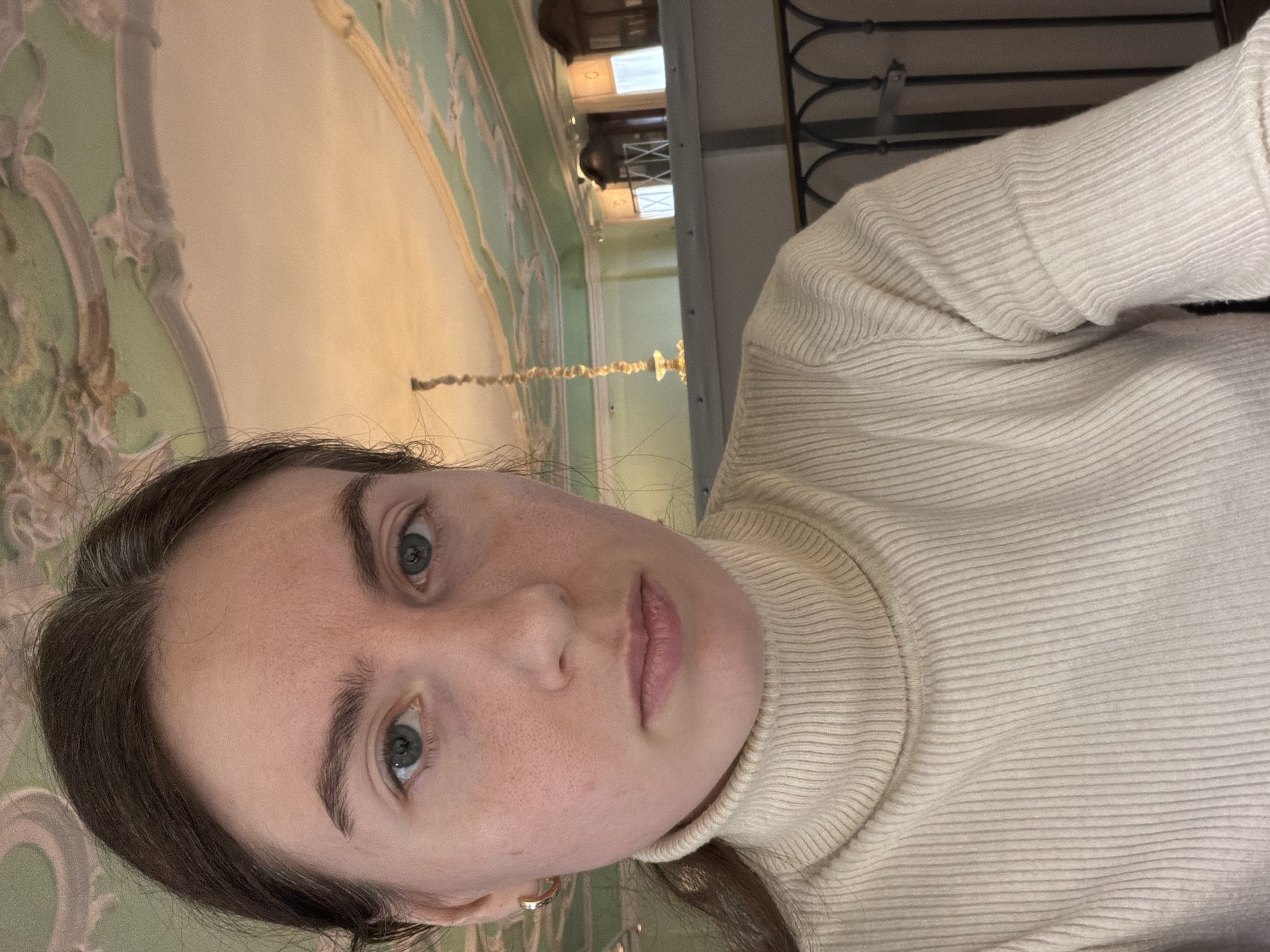 A young woman is the subject of a medium-shot photograph, taken indoors in Saint Petersburg, Russia. She faces the camera with a neutral expression. She has fair skin, light blue eyes, and dark brown hair. She is wearing a cream-colored turtleneck sweater. She is wearing earrings.
In the background is a building interior, with ornate, pale green and beige plasterwork visible on the walls and ceilings. A chandelier is visible. There is a metal railing and the upper part of a doorway.