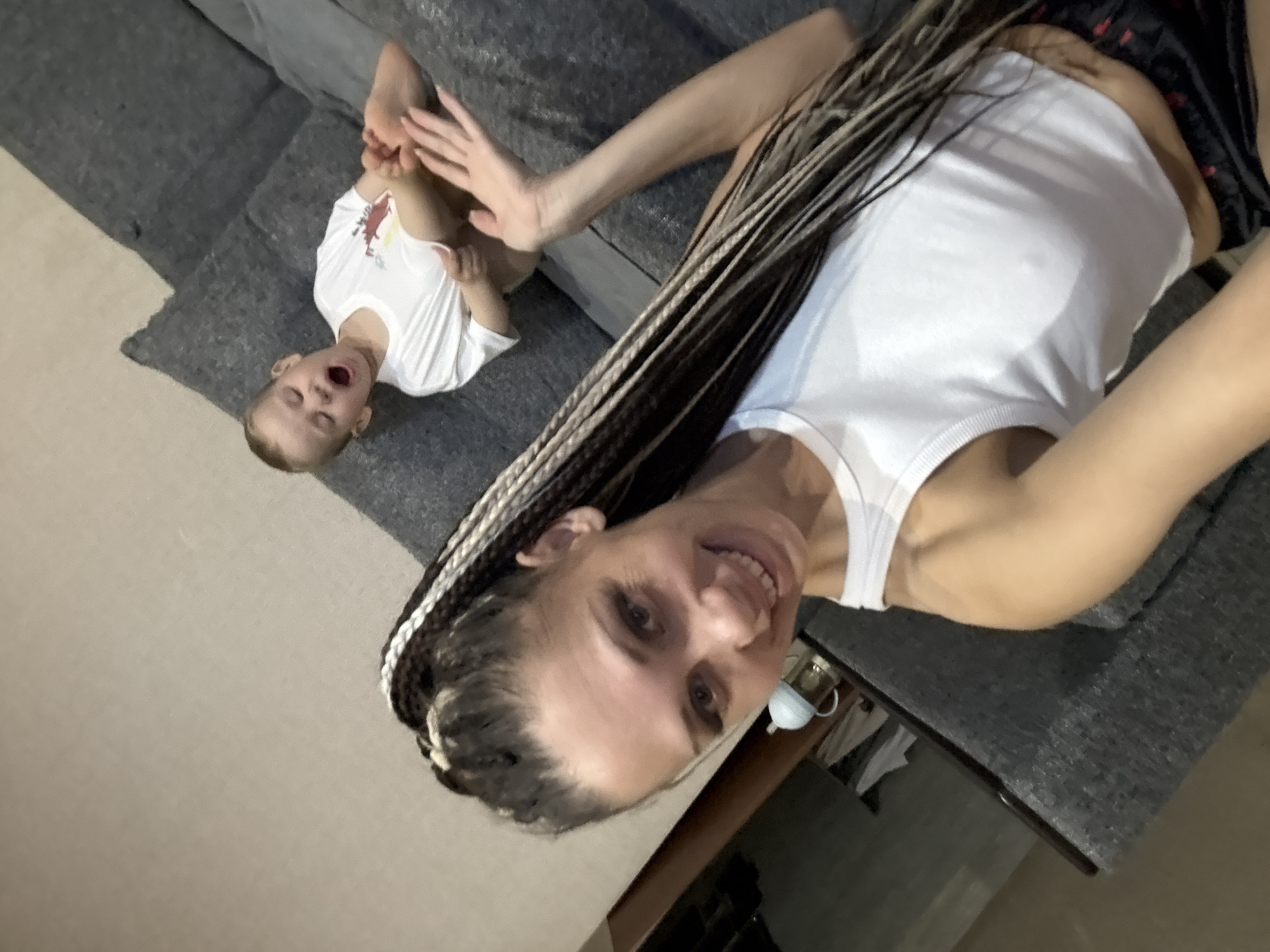 The image shows a woman with long braids, wearing a white tank top, smiling at the camera. To her left, a young child in a white shirt is lying on a gray sofa with their mouth open and hand raised towards their feet. The background includes a gray sofa and a room interior with neutral walls. The location mentioned, Rusanovo, Russia, cannot be verified from the image alone.
