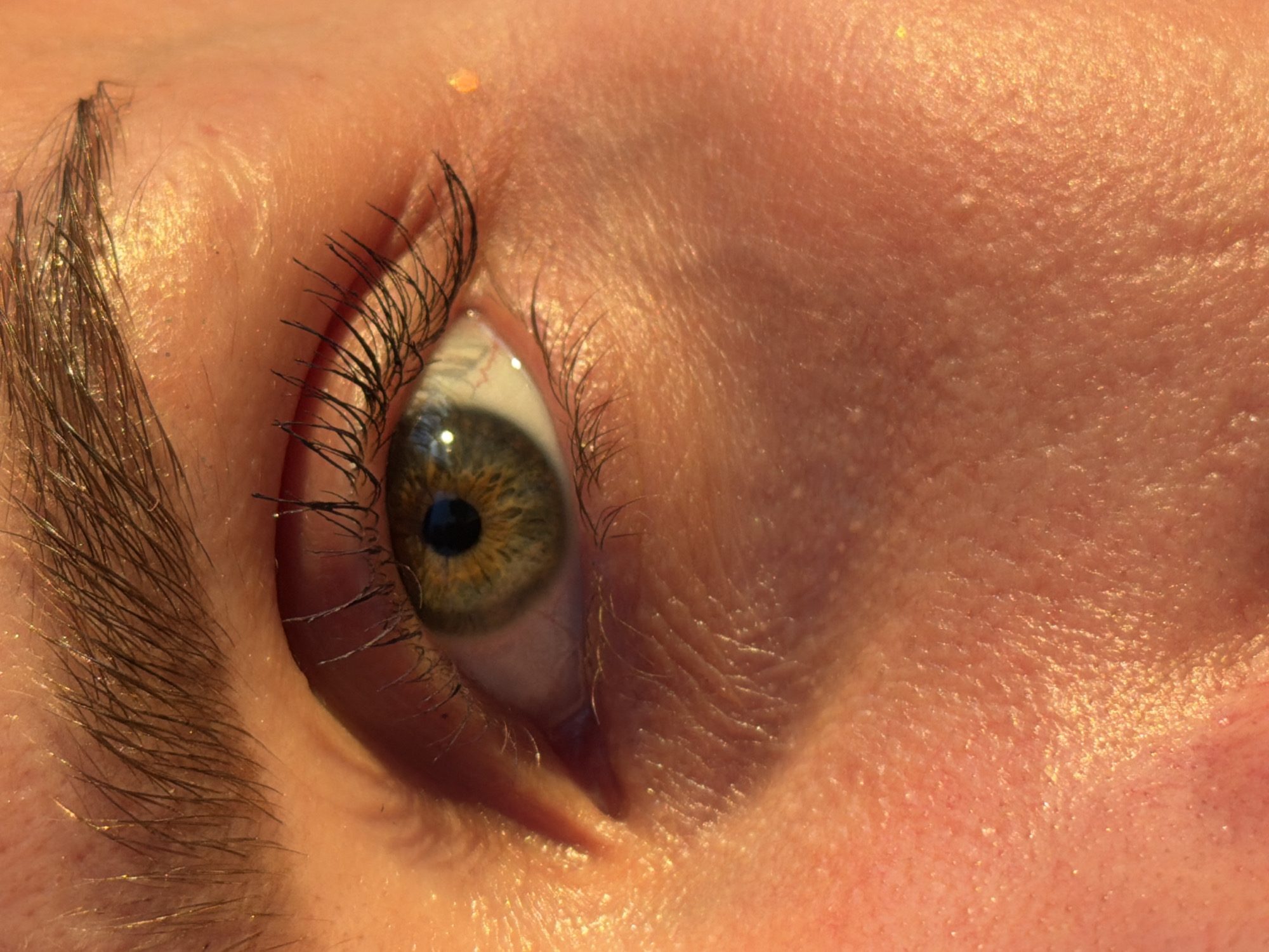 This is a close-up, macro photograph of a human eye. The eye is open and looking slightly to the left of the camera. The iris is a hazel green color with darker brown and yellow flecks. The pupil is dilated. Eyelashes frame the eye, and there are visible eyebrows above. The skin around the eye is illuminated by warm, golden light, casting shadows that highlight the texture of the skin, including fine wrinkles and pores. There are tiny flecks of glitter or shimmer on the skin, particularly on the cheekbone area. The background is out of focus, showing more of the skin and its texture. The image does not depict any specific location in the city of Chaikovsky, Russia, nor does it contain any identifiable objects or actions within that context.