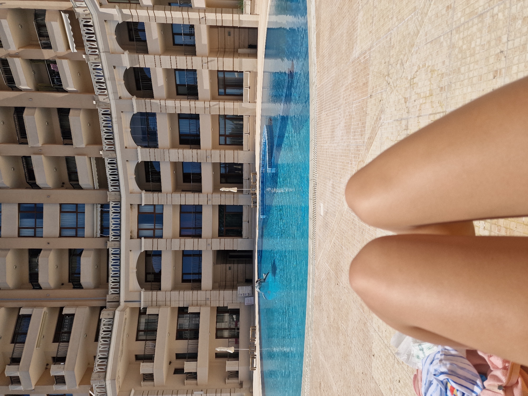 A first-person perspective shot shows a person's legs, clad in swimwear, lying on a tiled patio. Beyond the patio, a large, clear blue swimming pool is visible, with a person in mid-air, diving into the water. The background features a multi-story, cream-colored building with numerous balconies and windows.