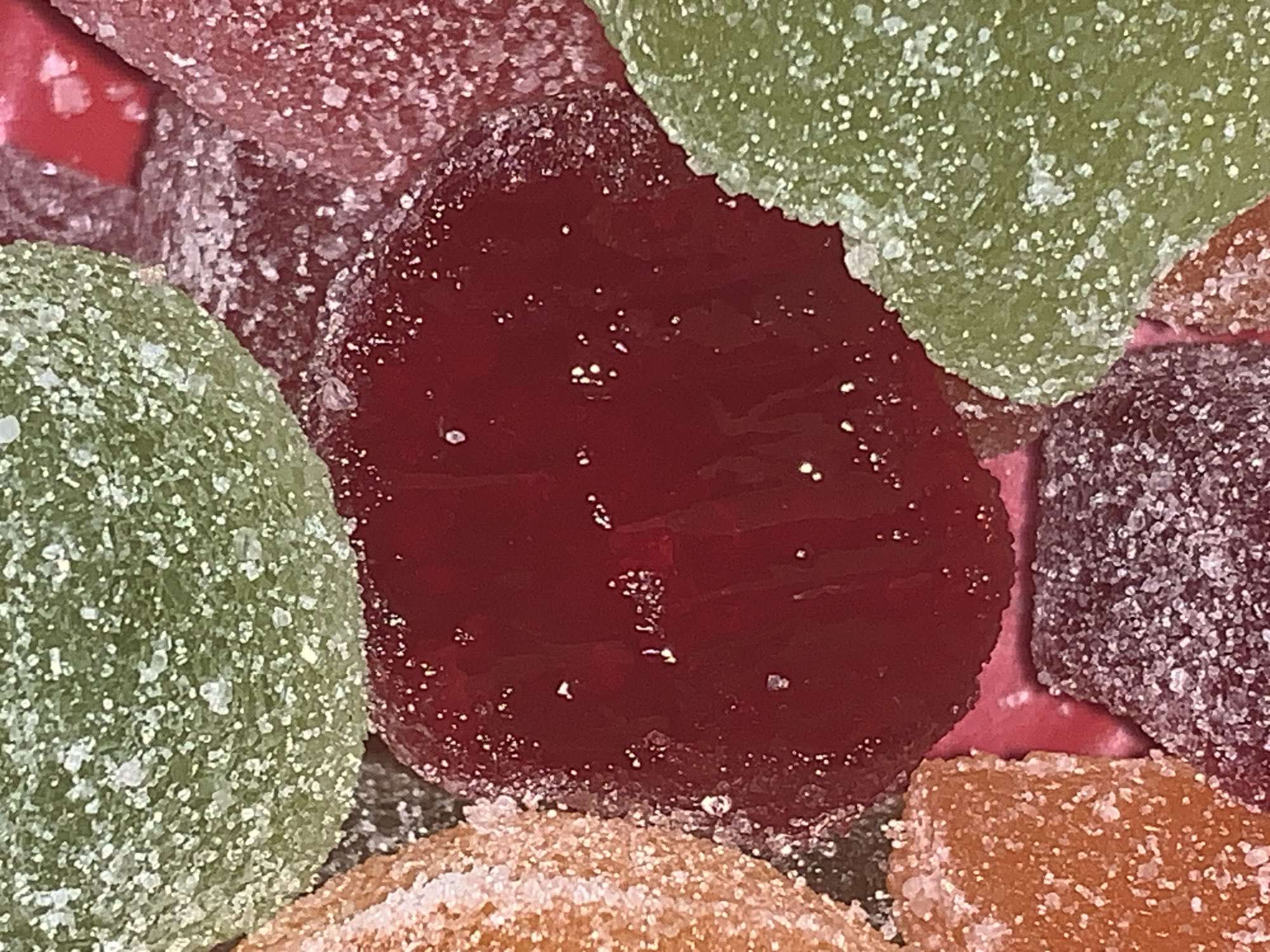This close-up image captures a vibrant assortment of sugar-coated gummy candies or fruit jellies, filling the entire frame. The primary subjects are several irregularly shaped confectionery pieces, each heavily dusted with white sugar crystals, creating a glittering, textured surface.
The candies display a range of rich, translucent colors. A large, prominent piece in the center is a deep, glossy red, its surface reflecting light with tiny sparkles. To its left, a bright, apple-green candy is visible, also coated in sugar. In the upper right, another green piece can be seen, partially overlapping with a reddish-pink background element. Towards the bottom and right, there are glimpses of other candies: one with a warm orange or amber hue, and another in a dark purple or maroon color.
The sugary coating is a notable detail, with individual crystals clearly visible, suggesting a rough yet appealing texture. The underlying jelly material appears soft and slightly gelatinous. The lighting is bright and direct, enhancing the candies' colors and making the sugar crystals sparkle. There are no discernible activities or events taking place, as it is a still-life arrangement of sweets. No people, visible text, or specific location cues are present. The image conveys a sense of sweetness and rich flavor through its vivid colors and inviting texture.
