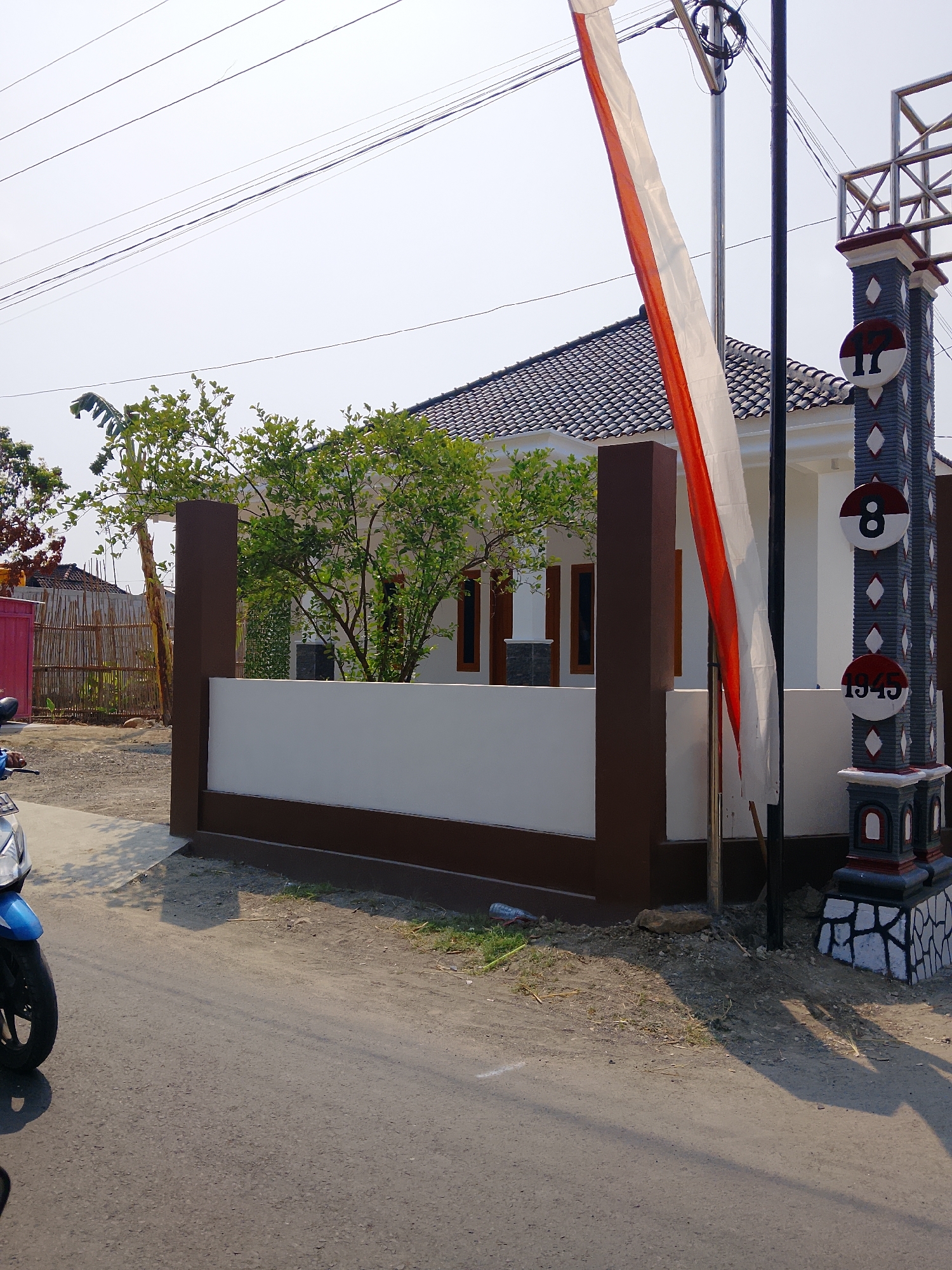 The image depicts a residential scene in Ngaglik, Indonesia. In the foreground, a paved road runs diagonally from the bottom left to the middle right of the frame. A blue and silver motorcycle is partially visible on the left side of the road, facing away from the camera. To the right of the road, a white and brown fence encloses a property. Behind the fence, a white house with a dark tiled roof is visible. Several green trees with leafy branches are situated within the property. A tall, dark pole stands to the right of the house, adorned with a vertical Indonesian flag, which is red and white. Adjacent to the pole is a decorative pillar with diamond patterns and circular emblems featuring the numbers "17", "8", and "1945", suggesting a connection to Indonesian Independence Day. The environment appears sunny with a clear sky. The background shows a bamboo fence and more vegetation, indicating a semi-urban or suburban setting.