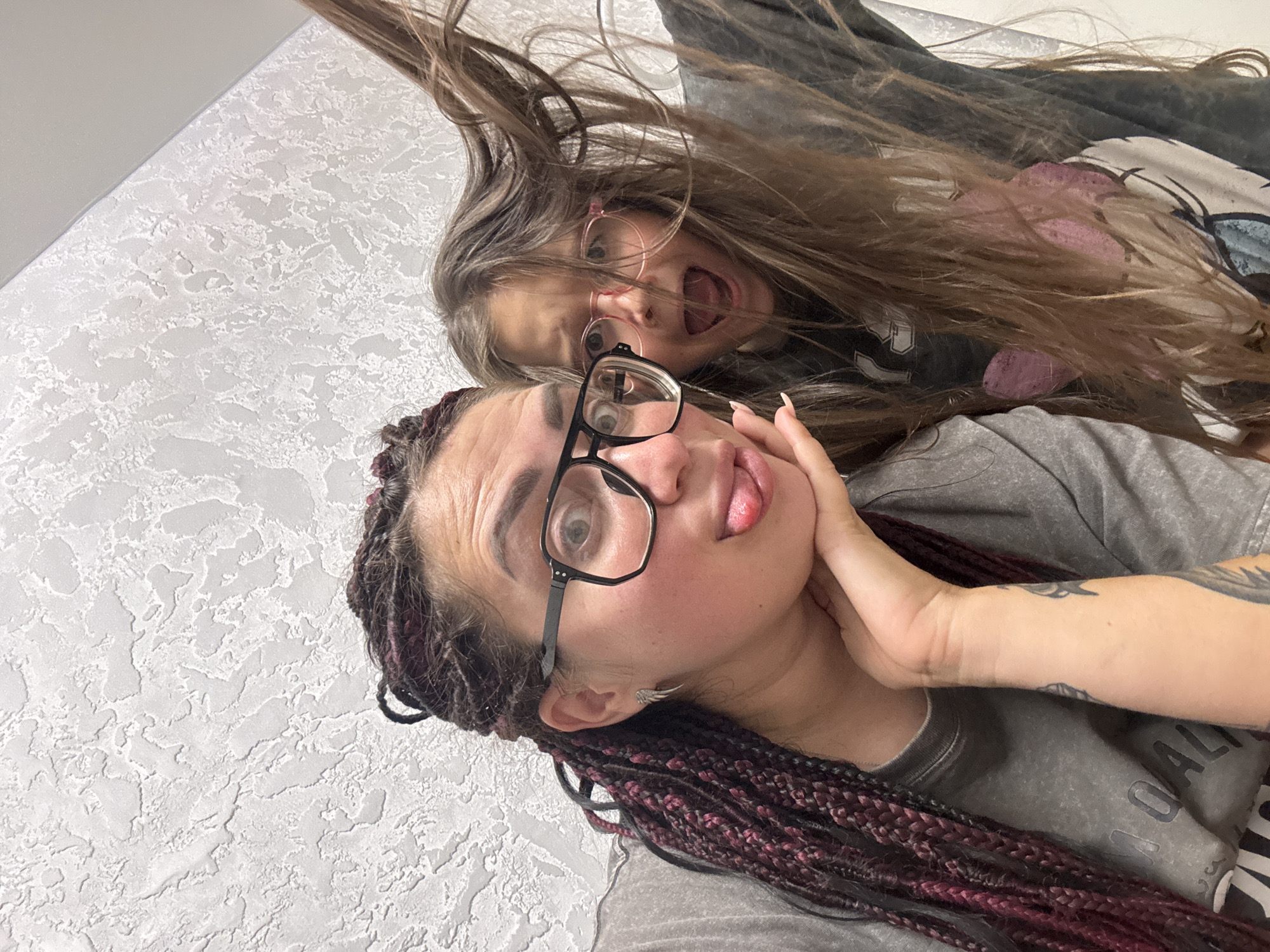 Two females are posing for a selfie against a textured white wall. The female in the foreground on the left, with braided dark hair with burgundy highlights, wears glasses and sticks out her tongue. Her left arm is propped on her chin, and she has tattoos visible on her arm. The female on the right, with long brown hair, also wears glasses and has her mouth open in a surprised expression. Her hair is flung forward, obscuring part of her face. The environment appears to be indoors. The image does not contain information to determine the location in the city of Tolyatti, Russia.