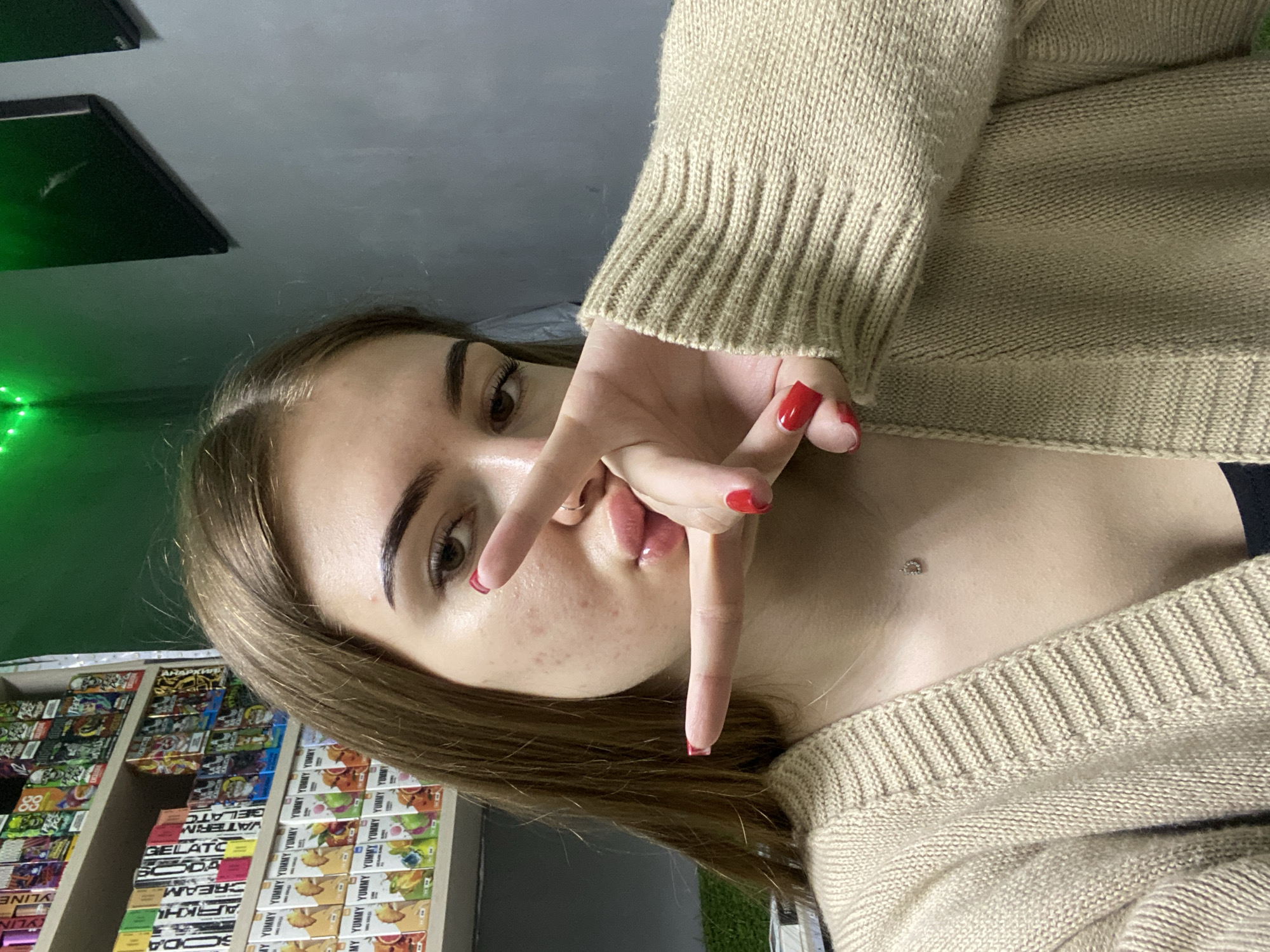 The image is a close-up portrait of a young woman, captured at an angle that implies a selfie. She has long brown hair, defined eyebrows, and brown eyes, looking directly at the viewer. Her right hand is raised, making a peace or victory sign with her index and middle fingers, partially covering her mouth. Her fingernails are painted bright red. She has some blemishes on her cheek and chin. A small, sparkling dermal piercing is visible on her upper chest.
She is wearing a light beige or tan knitted sweater with a visible ribbed texture on the cuffs and hem, and a darker garment underneath.
The background indicates an indoor commercial setting, possibly a convenience store or vape shop. To her left, shelves are densely stocked with numerous small, colorful, rectangular packages, likely disposable vape products. Green neon lighting is visible along the left edge of the frame. The ceiling above is gray with dark rectangular panels.
Visible text on the product packaging includes: "SODA", "CYLIN", "ARKHU", "CREAM", "YLINE", "ELATC", "VATERIM", "GELAT", "MY YUMMY YUMMY YUMMY YUMMY YUMMY YUMMY YUMMY", and "88". The repetitive "YUMMY" suggests flavor indicators on the products. The time of day is not discernible.