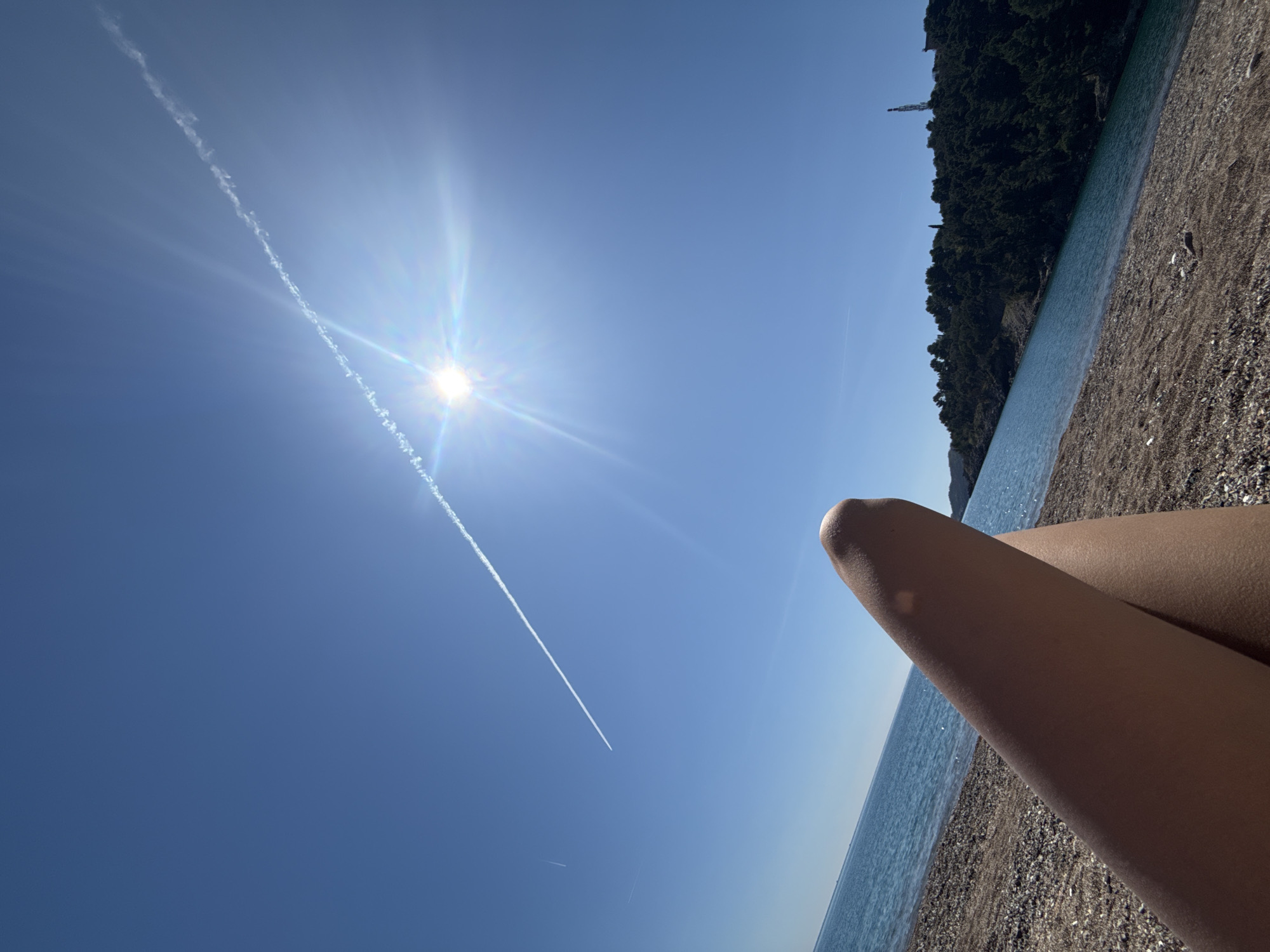 The image shows a sunny day at a beach. The sky is a clear blue with the sun shining brightly and a contrail visible. The beach appears to be pebbly, with the water's edge visible. Part of a person's legs is visible in the foreground, resting on the beach. In the distance, there's a stretch of land covered with green trees and shrubs. The city and location are not identifiable.