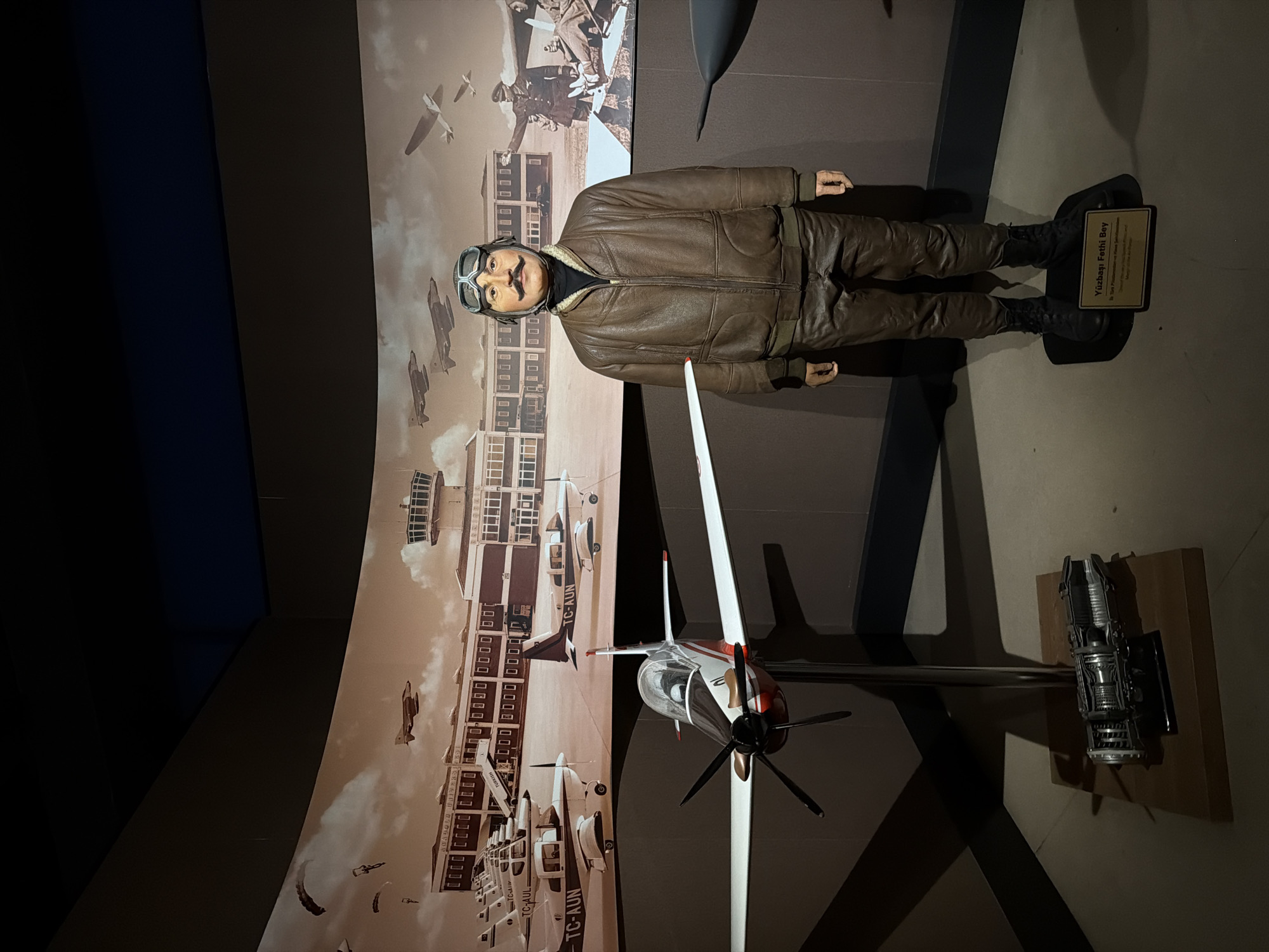 This media file depicts an aviation museum exhibit, likely located in Eskisehir, Turkey. The central subject is a life-sized mannequin of a male aviator, standing upright and dressed in historical flight attire. He wears a brown leather flight jacket, dark trousers, black boots, a dark turtleneck, and aviator goggles pushed up onto a cap, featuring a prominent mustache.
At the mannequin's feet, a small rectangular plaque reads "Yüzbaşı Fethi Bey," which translates to Captain Fethi Bey. Below this, there is smaller text in both Turkish and English, likely providing biographical details about the aviator, though it is not fully legible.
To the right of the mannequin, a scale model of a single-engine propeller airplane is displayed, predominantly white with red and black accents. Further to the right, a detailed miniature model of a jet engine component is mounted on a wooden base.
The background is dominated by a large, sepia-toned mural depicting an active historical airport scene. It features a grand airport building with a control tower, multiple vintage aircraft (including biplanes and monoplanes) on the tarmac and in flight, and several parachutists descending from the sky. Text like "TC-AUN," "TC-AUL," and "TURKISH" are visible on the aircraft and airport building within the mural, indicating Turkish aviation context. The scene evokes a sense of early 20th-century aviation history.