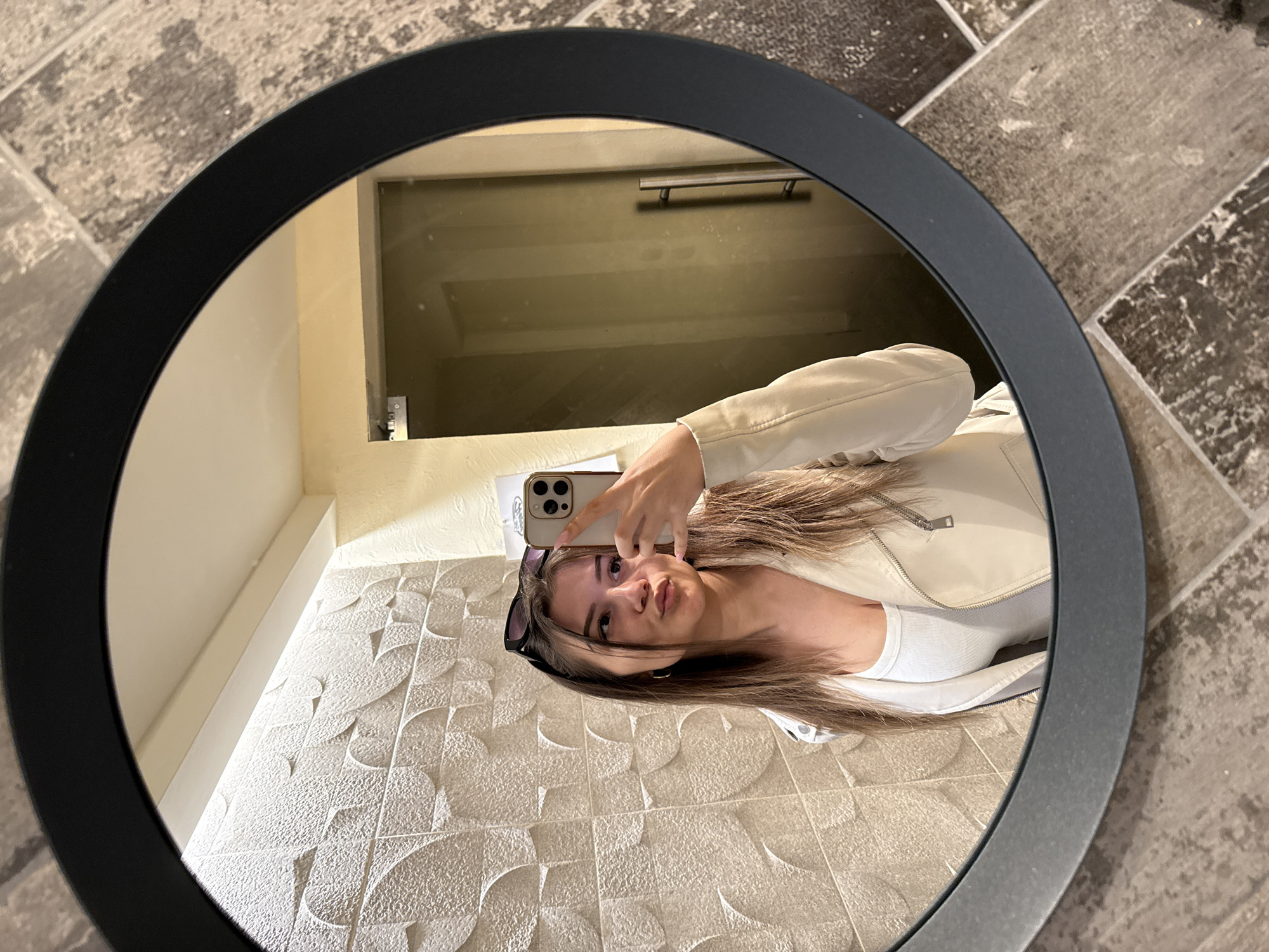 A young woman is taking a selfie in a round mirror. The mirror has a black frame and reflects the woman from the chest up. She is holding a phone in her right hand. She is wearing a white tank top and a white jacket, and sunglasses are atop her head. The reflection reveals that she is in a bathroom or restroom, with decorative white wall tiles behind her. The floor is tiled with gray and brown tiles. The location is possibly in Sofia, Bulgaria.