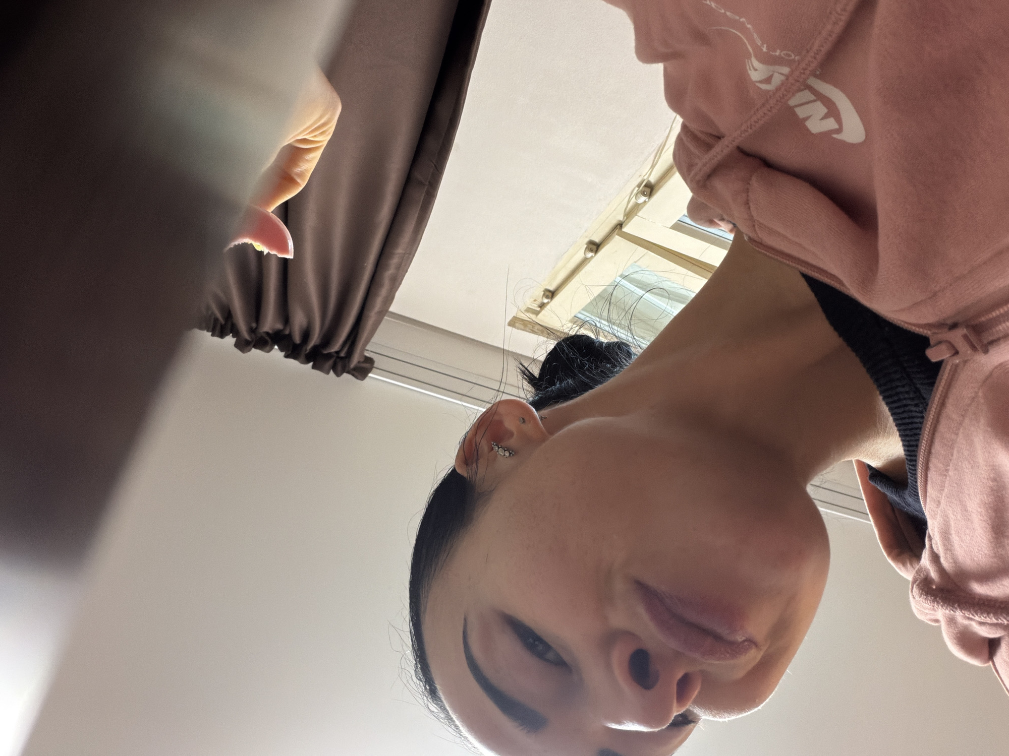 A low-angle perspective photograph shows a person, presumably female, looking down at the camera. The person has dark hair, dark eyebrows, and a fair complexion. She is wearing a light pink zip-up hoodie with a white logo partially visible on the sleeve. A black garment is visible underneath the hoodie at the neckline. The person has multiple piercings in her left ear. The background includes a window with a view of a light-colored wall and a curtain. It is not possible to verify the location of Saint Petersburg, Russia.