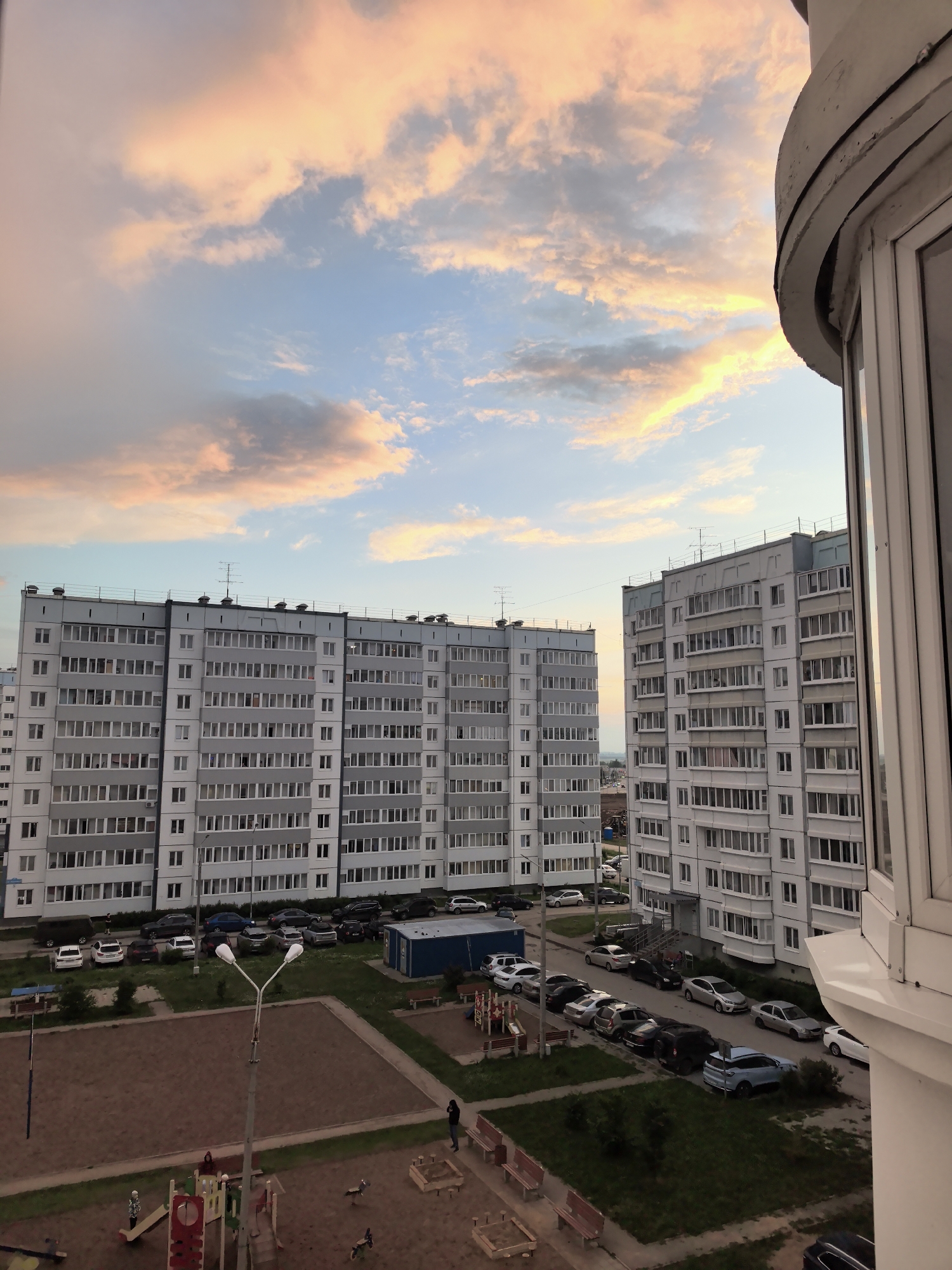 This image depicts a residential courtyard in Froli, Russia, during sunset. Two multi-story apartment buildings, constructed with light-colored facade panels and numerous windows, dominate the scene. The sky above is adorned with dramatic clouds painted in hues of pink, orange, and blue. In the foreground, a playground area is visible with swings, slides, and sandboxes. Several cars are parked in the designated parking areas between the buildings and the playground. A lamppost stands prominently in the courtyard. The overall environment suggests a typical urban residential area.