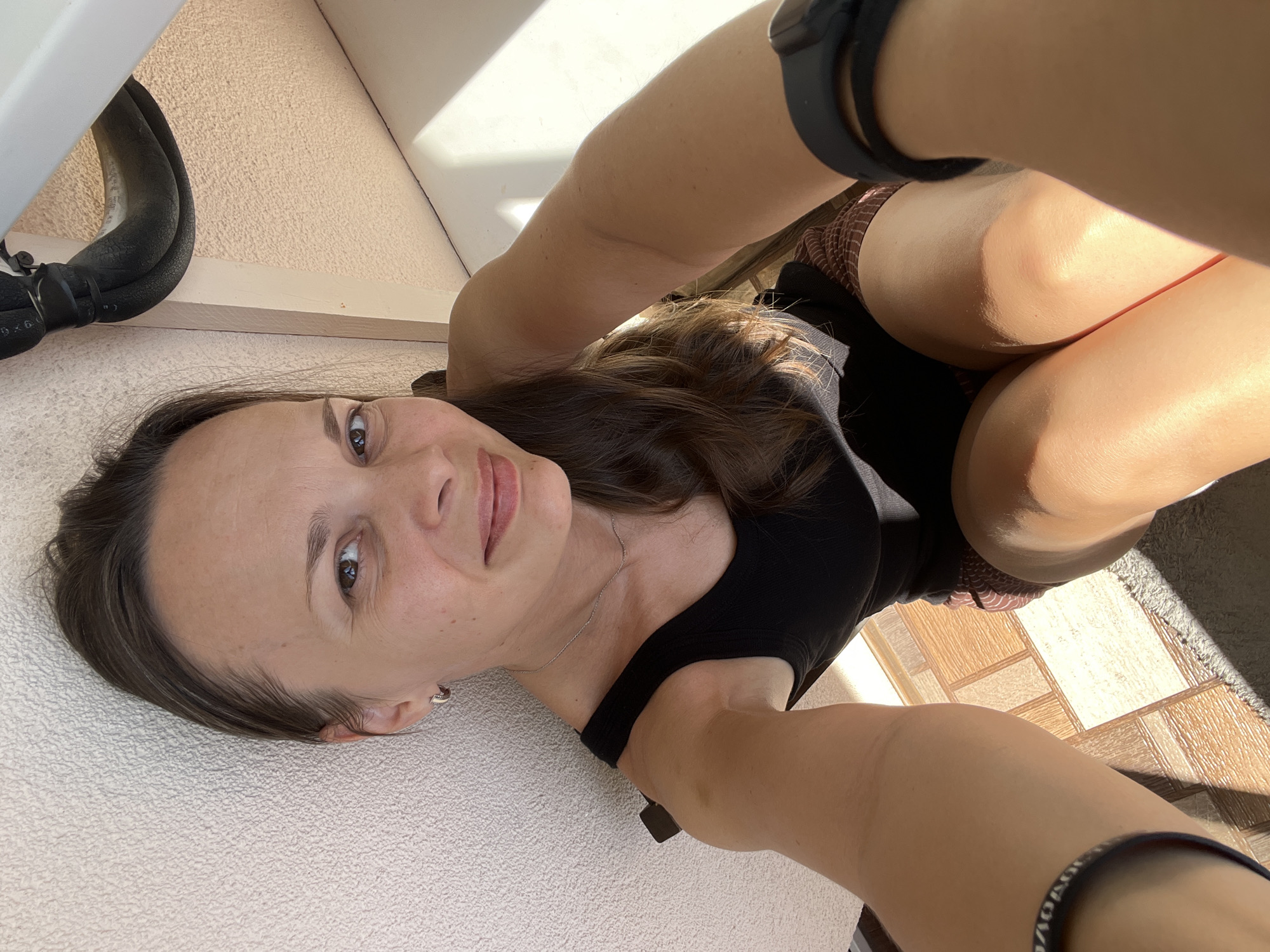 This media file captures a person taking a selfie, oriented sideways. The subject is a woman with long, brown hair, looking directly at the camera with a neutral expression. She is wearing a black tank top and what appears to be patterned shorts. A silver necklace is visible around her neck.
The setting seems to be outdoors, possibly on a balcony or patio, given the presence of outdoor tiling and sunlight casting shadows. The sunlight suggests it is daytime. In the background, there's a wall with a textured surface and what looks like part of an air conditioning unit or similar piping. The ground is covered in light-colored, textured material, possibly a rug or towel, and further back, there is tiled flooring.
On the left side of the frame, a black boot is visible, suggesting the person might be wearing boots or shoes. On the right arm of the subject, a black smartwatch or fitness tracker is worn. Another dark band with white text is visible on the right forearm. The overall mood is casual and relaxed.
Visible text includes "9x6" on a piece of black tape attached to the piping, and some unreadable text on the band on the right forearm. The location context is Tolyatti, Russia.