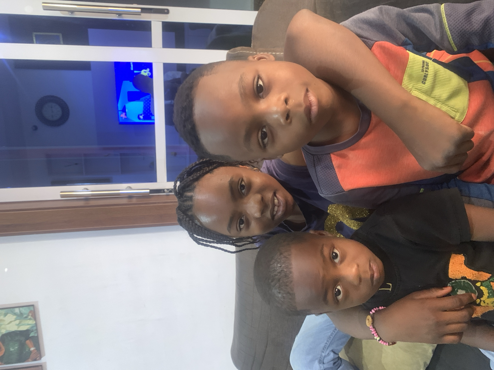 Three children are closely grouped together in an indoor setting. The topmost child wears a blue and orange shirt, while the child in the middle has braided hair and a navy shirt. The bottom child wears a black shirt with a colorful graphic and a pink bracelet. A window is visible in the background with a clock and television reflected in the glass. The location in the city of Jos, Nigeria, cannot be determined from the image.