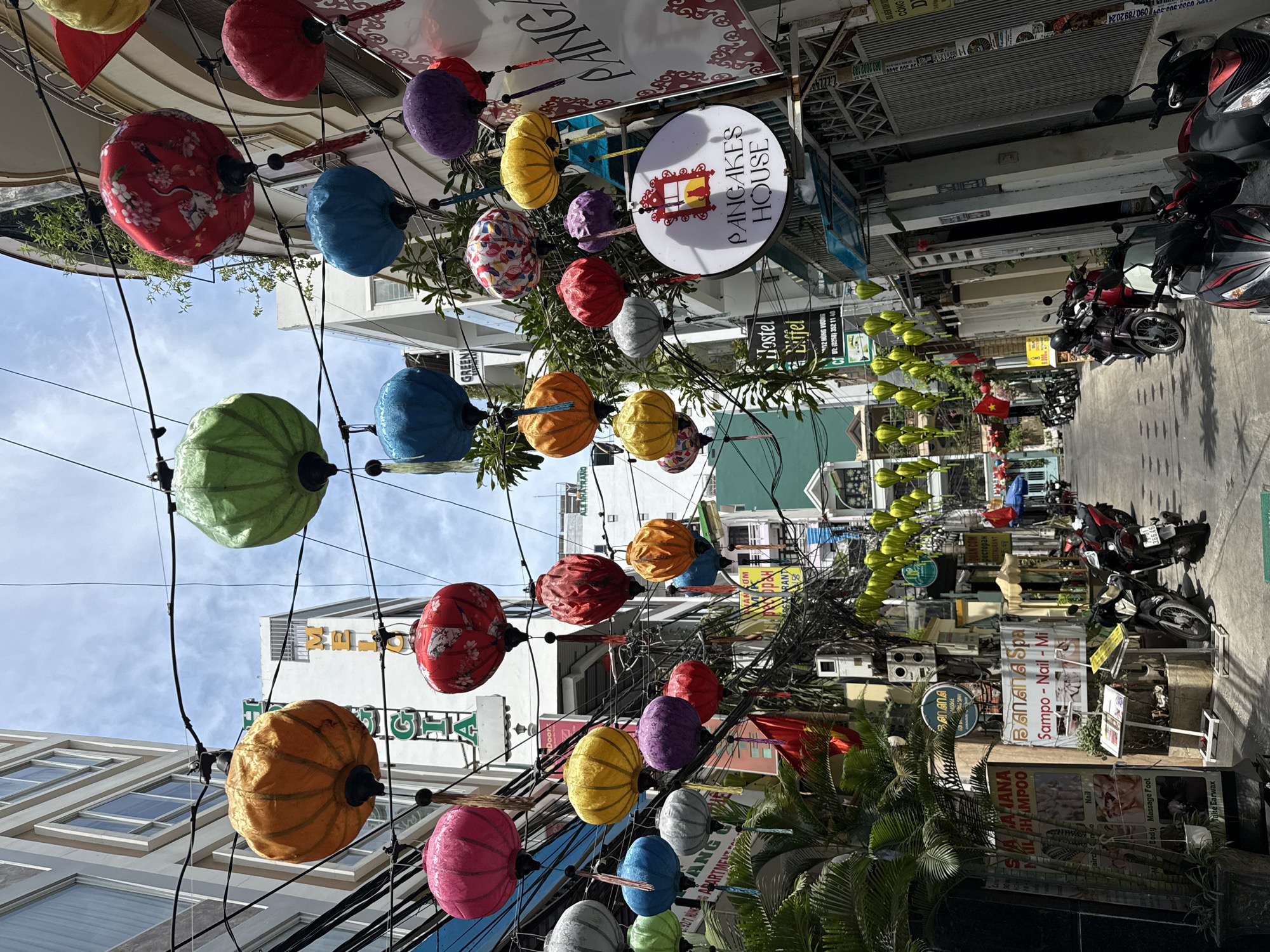 The image shows a street scene in Nha Trang, Vietnam, with a colorful display of lanterns hanging above. On the right side of the street, parked motorcycles and signs for businesses like "Banana Spa" and "Pancakes House" are visible. The buildings along the street are varied in style and height, with some showing signs of aging and others appearing more modern. The sky is partially visible through the gaps between buildings and lanterns, with patches of blue and some clouds. Overhead, tangled power lines run alongside the lantern strings.