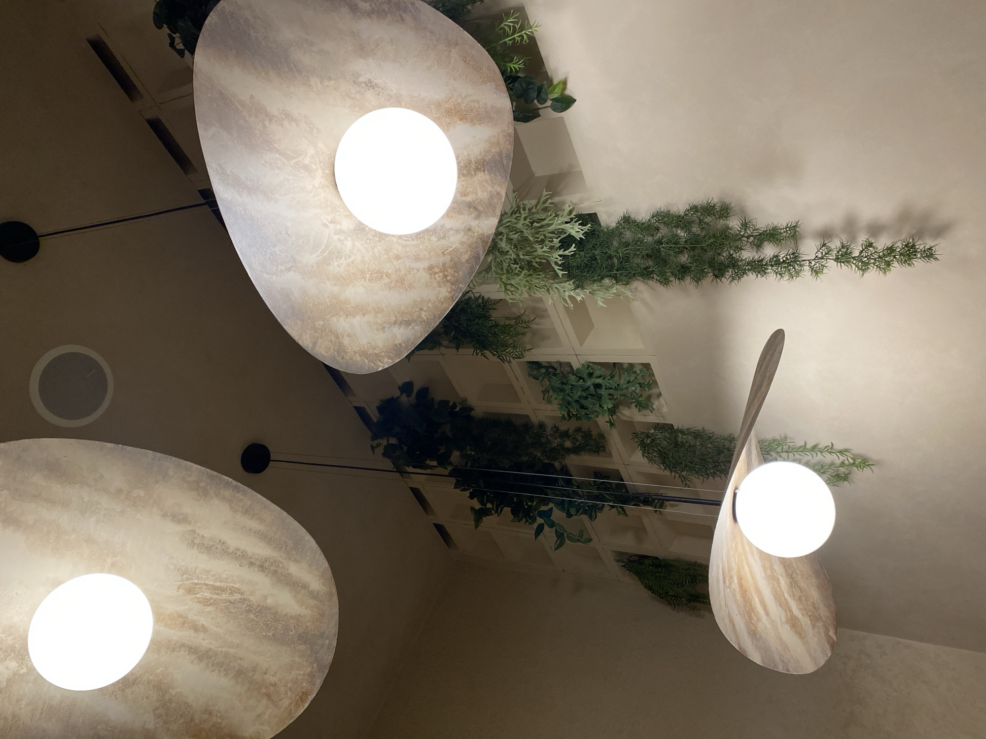 A low-angle, indoor shot depicts three modern light fixtures hanging from a ceiling. Each fixture has a circular, marbled-patterned lampshade with a bright, round bulb at its center. The ceiling features a decorative trellis pattern with green plants arranged within the squares of the trellis. A small, circular object, possibly a speaker, is visible near the upper left of the frame. The overall lighting is warm, and the perspective is angled upwards. There is no information provided within the image to associate it with Izhevsk, Russia.