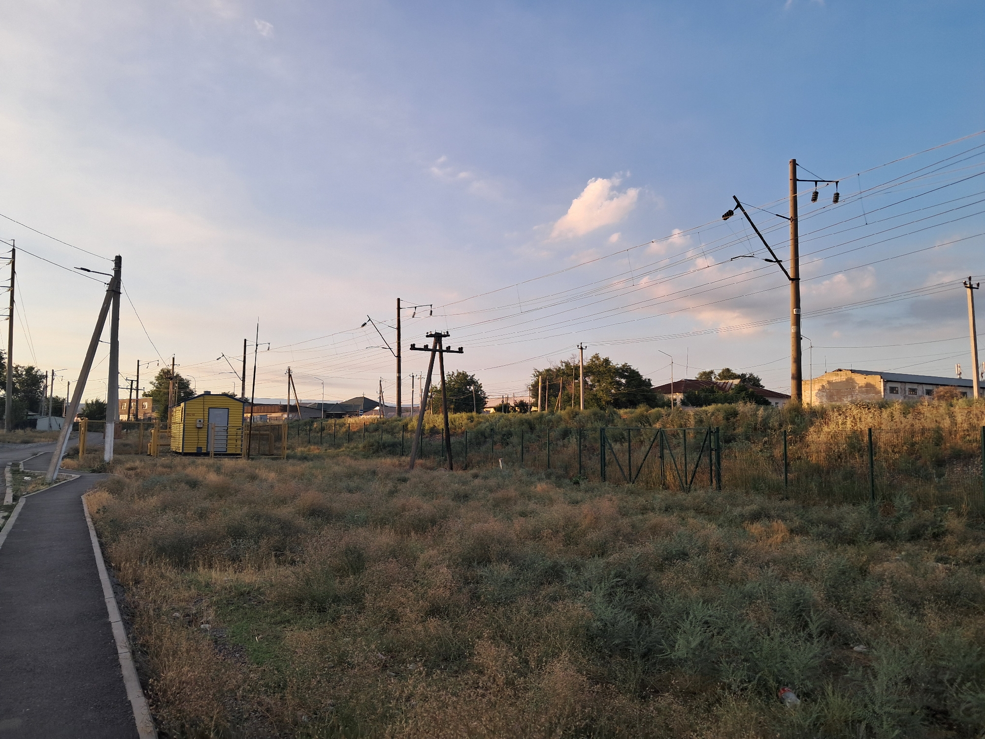 This media file depicts an outdoor scene in Shymkent, Kazakhstan, likely during late afternoon or early evening, as indicated by the warm, soft lighting and long shadows. The setting appears to be an undeveloped or rural area adjacent to infrastructure.
The primary subjects are utility poles carrying numerous electrical wires, which traverse the sky. In the foreground, a paved pathway curves to the left, bordered by a curb. To the right of the pathway, a patch of overgrown, dry grass and vegetation fills the mid-ground, separated by a green chain-link fence from a small, bright yellow building with a single door and what appears to be a window or vent. Beyond the fence and yellow building, several other structures, possibly industrial or residential, are visible in the distance, as are more utility poles and trees. The sky is a clear blue with a few wispy clouds, one of which is tinged with pink, further suggesting the time of day.
There are no people visible in the image. The activity taking place is passive, with the scene focused on the infrastructure and the natural landscape. Notable details include the abundance of electrical infrastructure, the dry, wild vegetation, and the isolated yellow building. No discernible text is visible in the image. The overall mood is tranquil and somewhat stark, typical of transitional or undeveloped urban fringe areas.