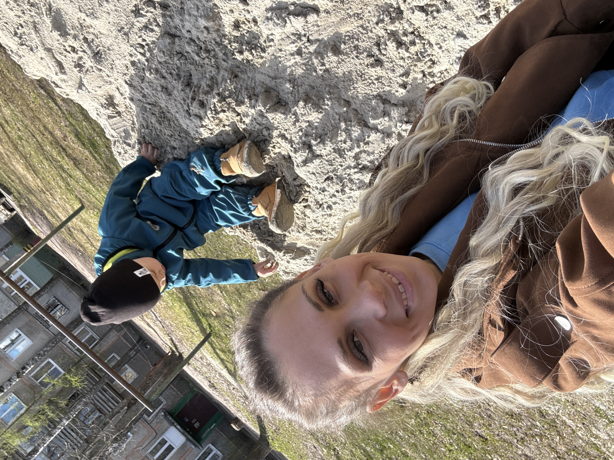The image captures a woman and a child outdoors. The woman, with blonde curly hair, is wearing a brown jacket over a blue top and is smiling towards the camera. Next to her is a child wearing a blue jacket, black hat, and brown shoes, sitting on sandy ground. In the background, there's a building with windows and a grassy area, indicative of an urban environment. Based on the architectural style of the building, the picture could plausibly be from Rusanovo, Russia. The interaction is a candid moment of a woman and a child outside.