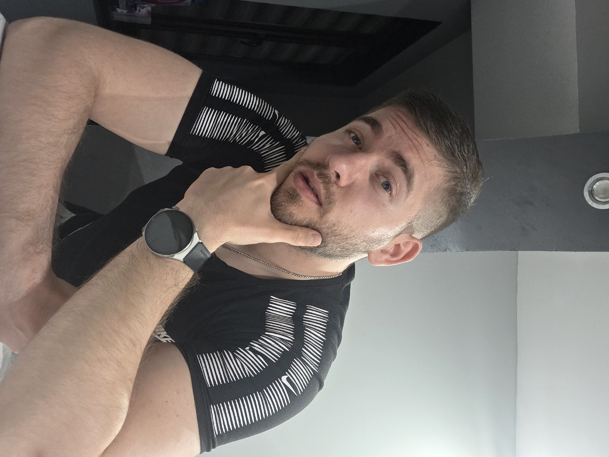 A man is looking at the camera with his hand on his chin. He has short brown hair and a light beard. He is wearing a black t-shirt with white stripes, and a silver watch on his left wrist. He is wearing a silver necklace. The background consists of a gray wall and a white wall. The location is unknown and cannot be determined to be in Yagodovo, Bulgaria based on the image.