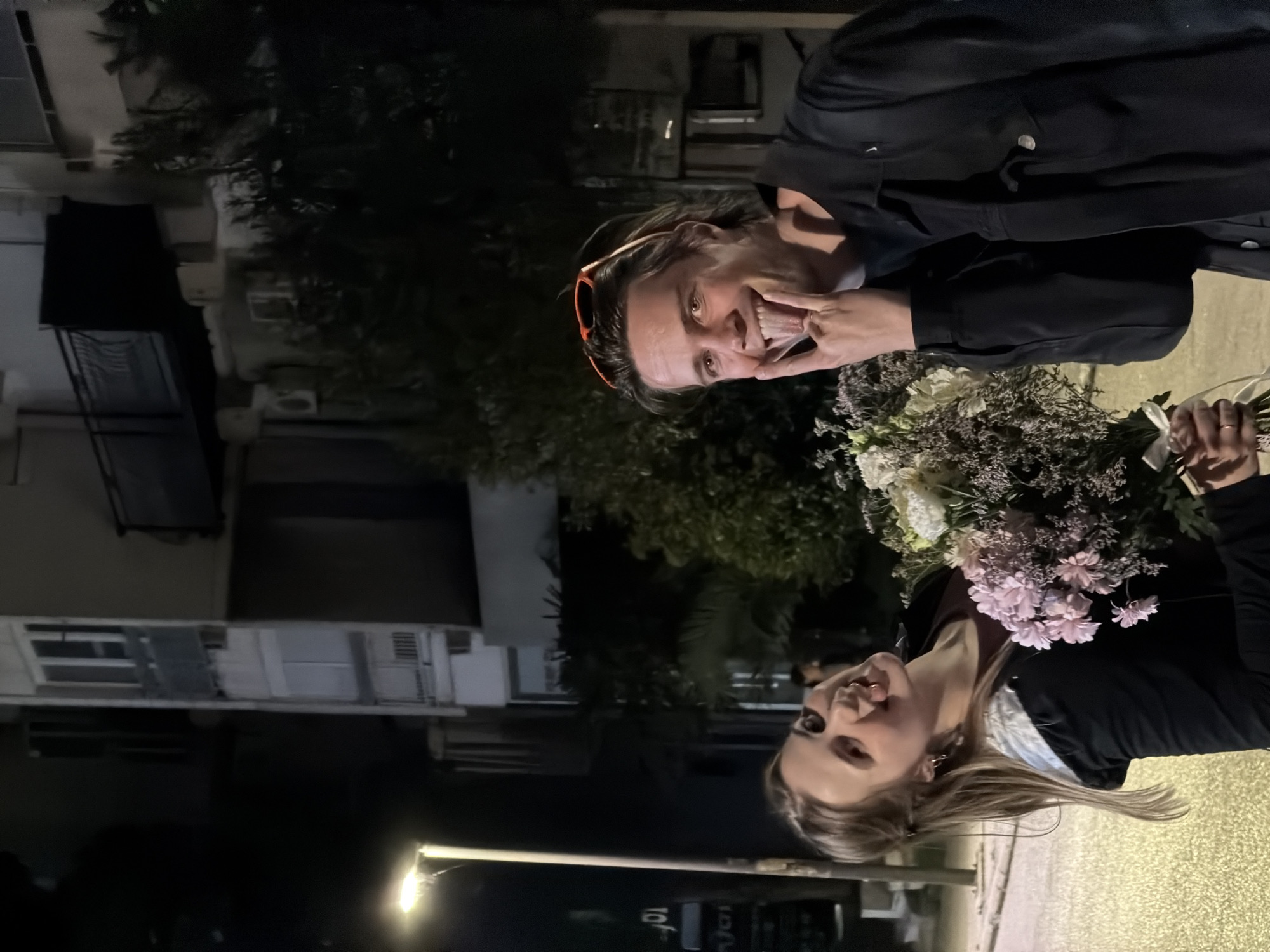 The image shows two women posing for a photograph outdoors at night. The setting appears to be an urban or suburban street with buildings and trees in the background, illuminated by artificial light.
The woman on the left has dark hair, with orange-framed sunglasses pushed up onto her head. She is wearing a dark jacket and makes a playful face at the camera, pulling at the corners of her mouth with her fingers and sticking out her tongue, revealing a cheerful expression.
The woman on the right has light, blonde hair and is also wearing a dark jacket over what appears to be a reddish top. She is smiling broadly towards the camera. She holds a large, mixed bouquet of flowers containing white, light pink (possibly chrysanthemums), and purple or dried floral elements. Her hands also hold a smaller bunch of white flowers tied with a white ribbon, partially visible behind the main bouquet.
The background is dark, signifying nighttime. Visible elements include multi-story buildings with windows and what appear to be air conditioning units, and dark trees or bushes. A bright street light or similar light source is visible on the lower left side of the frame, and near it, a partial sign displaying "10f" can be seen. The overall mood is happy and candid.
