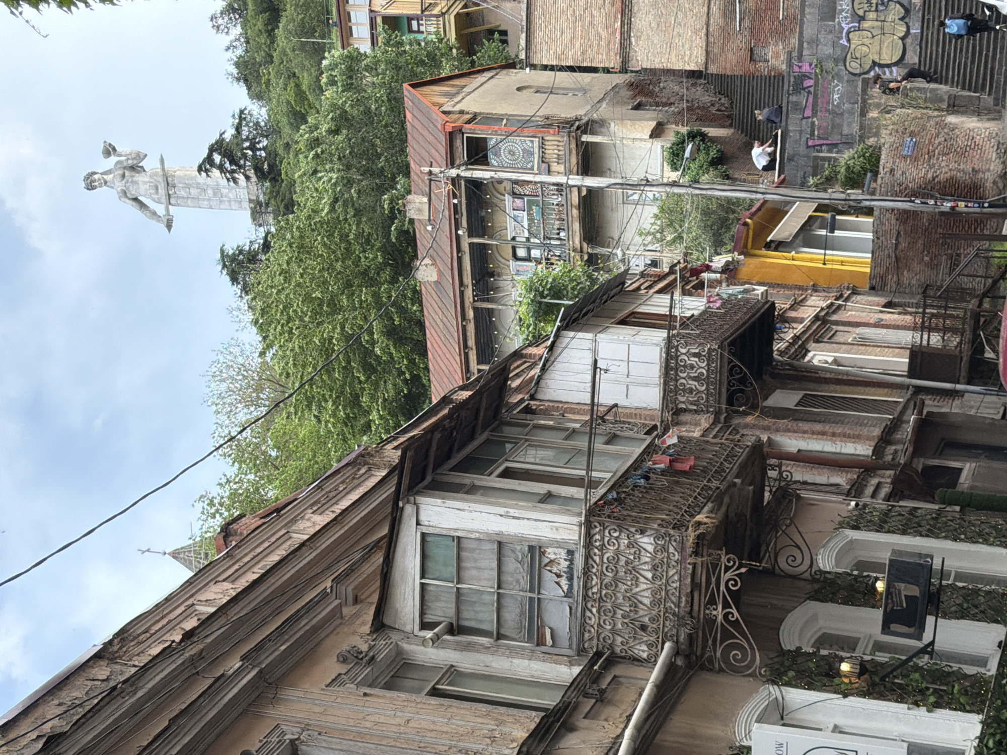 The image shows a view of the old town in Tbilisi, Georgia. In the background on a hill, the statue "Mother of Georgia" (Kartlis Deda) is visible. The foreground consists of old, dilapidated buildings with ornate balconies. There is a stone stairway to the right. Graffiti is visible on the stairway wall. People are on the stairway and the rooftops. Electrical wires run across the scene. A restaurant or shop is visible on the ground floor.