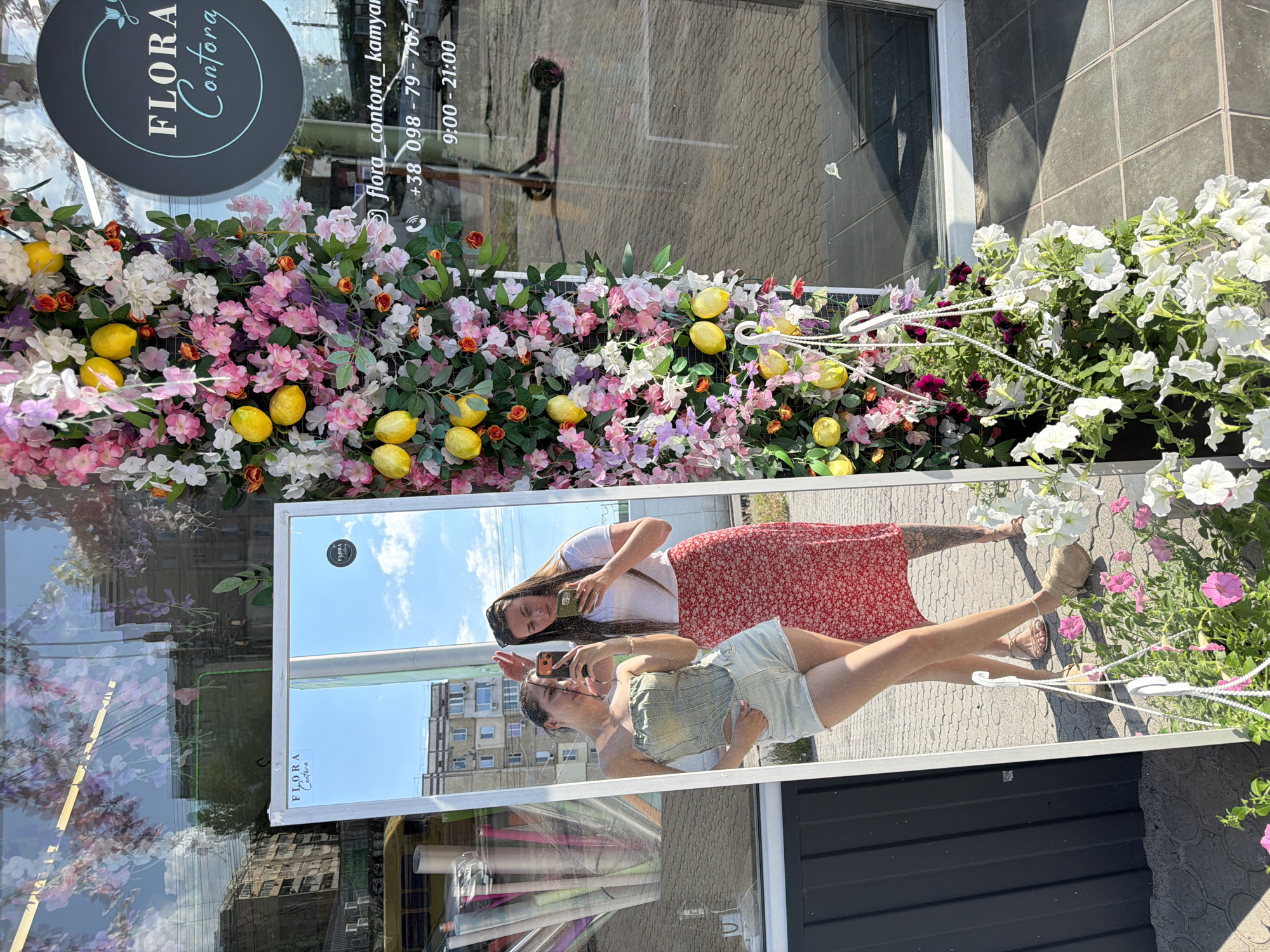 In the city of Unknown, Unknown, two young women are posing for a mirror selfie in front of a flower shop. The woman on the left is wearing a light green strapless top and light blue denim shorts, holding her phone with her left hand and resting her right hand on her hip. The woman on the right is wearing a white t-shirt and a red floral print skirt, holding her phone with her right hand and wearing sunglasses on her head. The flower shop has a large display of artificial flowers and lemons hanging on the right side of the image. A large mirror is positioned in front of the shop, reflecting the two women and the sky. In the reflection, the buildings across the street are visible. A black electric scooter is parked near the shop's entrance, and a portion of the shop's interior is visible through the glass door. The ground is paved with gray tiles.