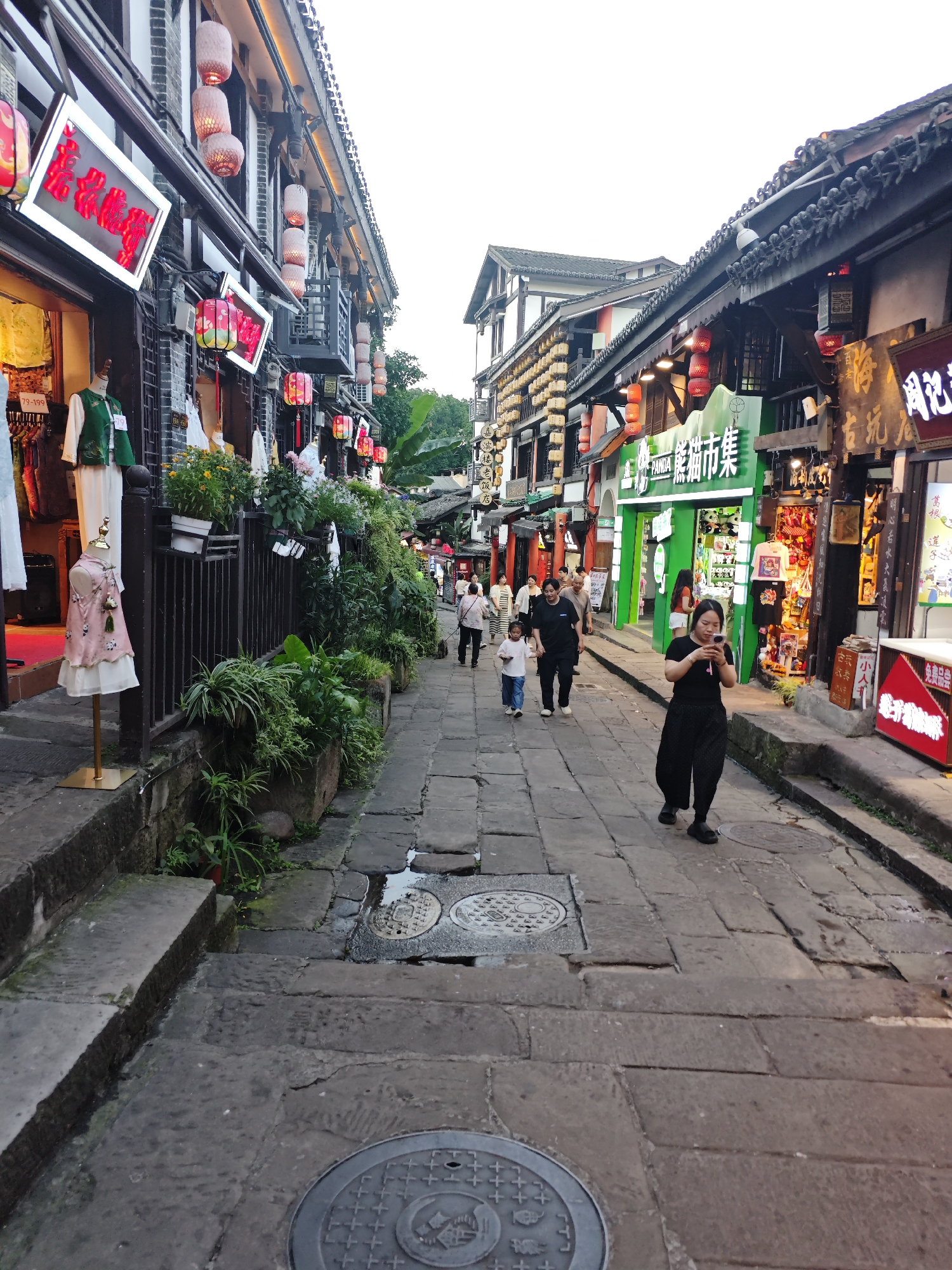 A narrow stone-paved pedestrian street in Shapingba District, China, is lined with two-story buildings with traditional Chinese architectural elements. The buildings feature shops with signs in Chinese and decorations like red lanterns. On the left, a clothing store displays mannequins in traditional outfits. Plants adorn the fences and ledges of the buildings. Several people walk along the street, including a woman in black pants and shirt looking at her phone, a woman holding the hand of a child, and other pedestrians in the distance. The sky is overcast. Manhole covers are visible on the stone pavement.