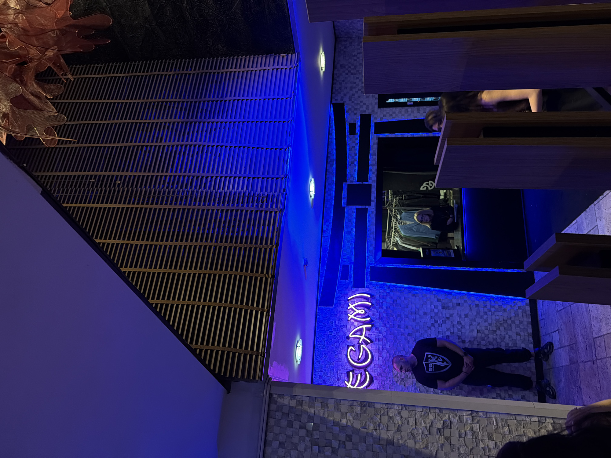 The image depicts the exterior of a shop called "EGAMI" in Sofia, Bulgaria. The facade features a section of dark-colored latticework, illuminated with blue light. Below the latticework, a neon sign spells out "EGAMI" in white. A man wearing a black shirt stands near the entrance. He appears to be looking down. Parts of other individuals are visible, including a person inside the shop and another near the edge of the image. The wall is decorated with light-colored tiling. Some wooden shelving or structures are visible along the right side of the frame.