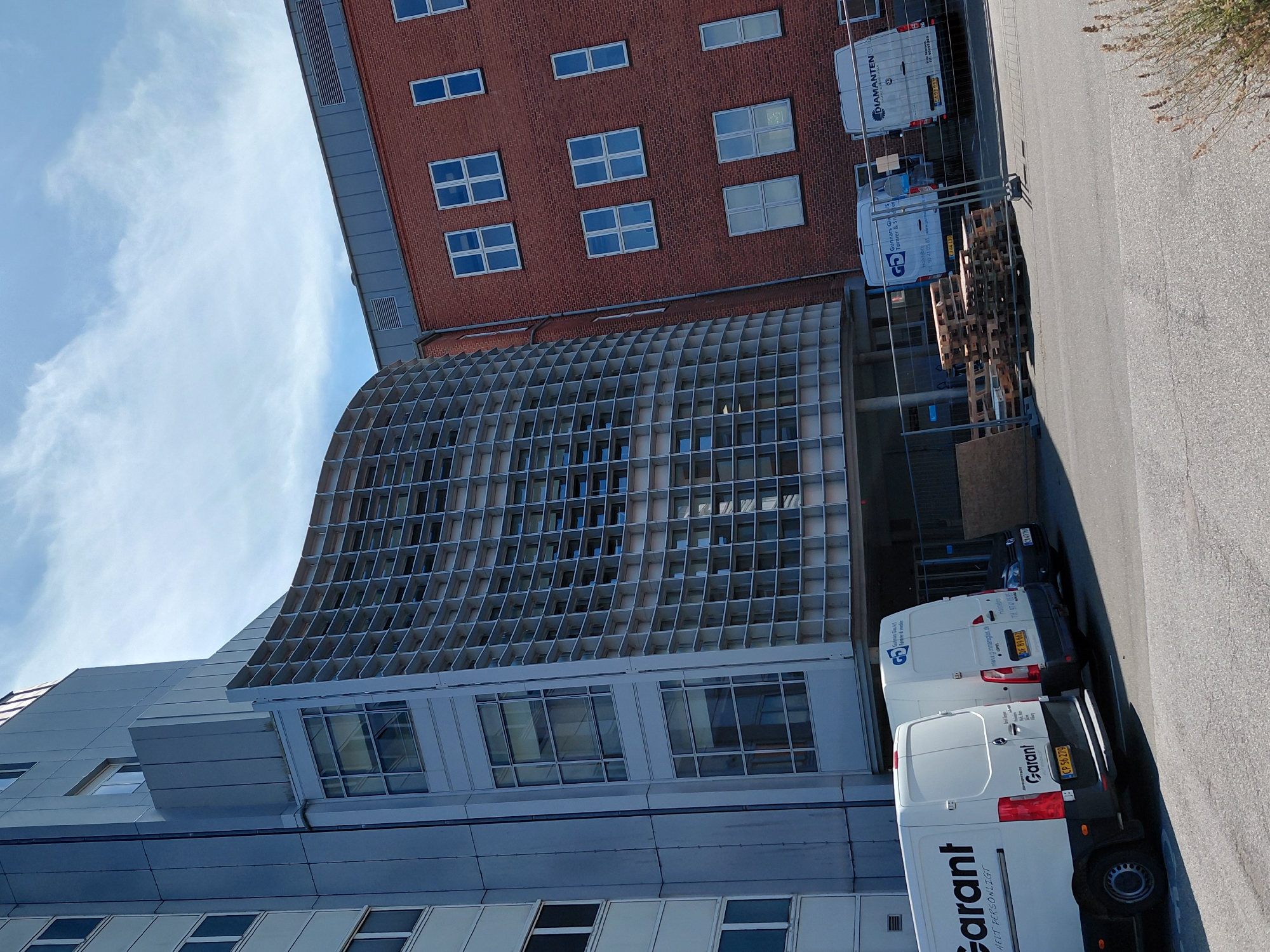 The image shows the exterior of a modern building with a curved facade covered in a grid-like structure, adjacent to a red brick building. Several white vans are parked in front of the buildings. On the left, a white van with "Garant" written on its side is parked. To its right, another white van with "Garant" and "Gunnars Glas A/S" logo is visible, along with the text "Holstebro Tlf. 97 41 05 85 www.gunnarsglas.dk". Further to the right, a third white van with "DIAMANTEN" logo and "CK 02 010" license plate is parked. The background shows a blue sky with scattered clouds. The overall scene appears to be an industrial or commercial area.