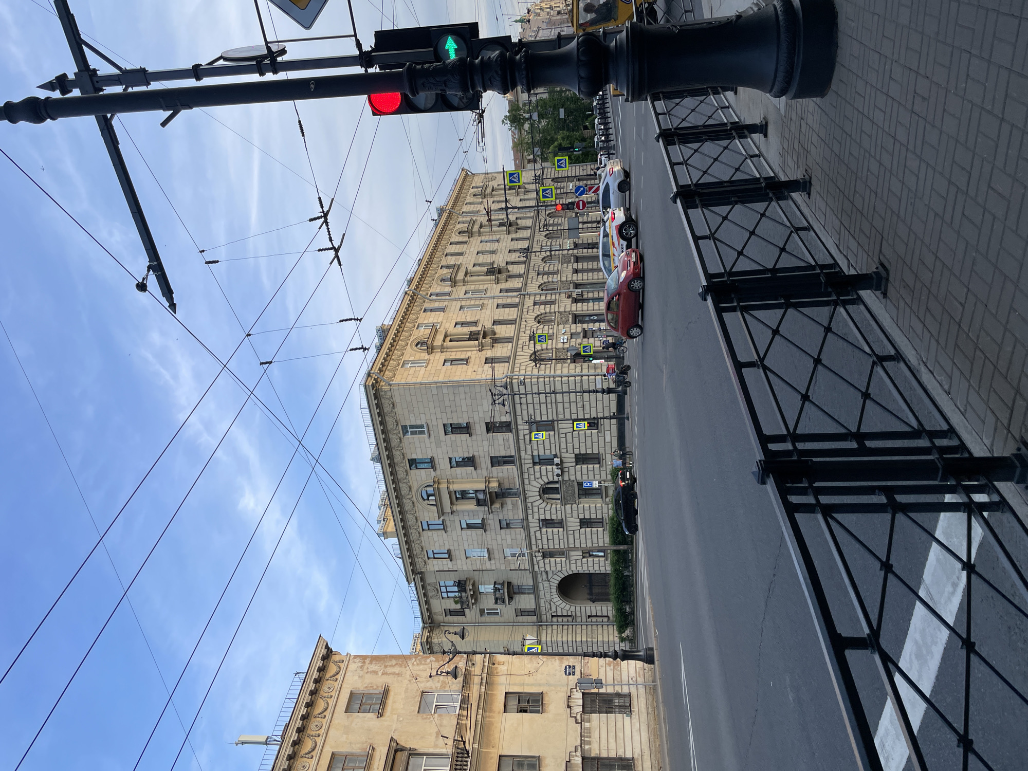 This image captures an urban street scene at the intersection of Moskovsky Prospekt and Gastello Street in Saint Petersburg, Russia, specifically focusing on the area around Moskovsky Prospekt 184 (left) and 186 (right). The time of day is late afternoon or early evening, evidenced by the soft, directional light and clear blue sky with sparse, wispy white clouds. In the foreground, a dark, decorative metal fence with a diamond lattice pattern runs parallel to the street, separating the asphalt road from a sidewalk paved with dark brick pavers. A large, black, ornate cast-iron streetlamp base is partially visible on the far right. A white pedestrian crossing is delineated on the asphalt directly in front of the fence. The midground features a wide, multi-lane asphalt road with white lane markings. Vehicular traffic is present: a red compact car is positioned in the rightmost lane, possibly waiting to turn or proceed. Behind it is a white SUV, identified as a Yandex.Taxi vehicle by yellow and black branding. Further back, another light-colored SUV is visible. On the far left lane, a black sedan is present. A yellow utility or maintenance vehicle is partially visible on the far right. Multiple traffic lights are active; the pole in the foreground displays a solid red circular light and an illuminated green arrow indicating a right turn. Several yellow diamond-shaped pedestrian crossing signs with black pedestrian figures are mounted on poles along the streets. A blue rectangular sign with a white 'P' (parking) is affixed to the main building on the right. The background is dominated by two multi-story buildings. The building on the left (Moskovsky Prospekt 184) is an older structure with a light-brown/ochre facade, showing signs of age and wear. It features multiple windows and subtle decorative elements. In front of it, another black ornate streetlamp stands. The main building on the right (Moskovsky Prospekt 186) is a large, imposing structure in a light-beige/grey color, exhibiting Stalinist Neoclassical architectural style. It features a prominent arched entrance at ground level, secured by a dark metal gate, numerous windows, and multiple small balconies with black metal railings. Decorative cornices, pilasters, and other classical details adorn its facade. Green bushes are planted along its base. Beyond these buildings, the green canopy of trees and other distant structures are visible under the clear sky. Overhead, an extensive network of black electrical wires, characteristic of tram or trolleybus systems, crisscrosses