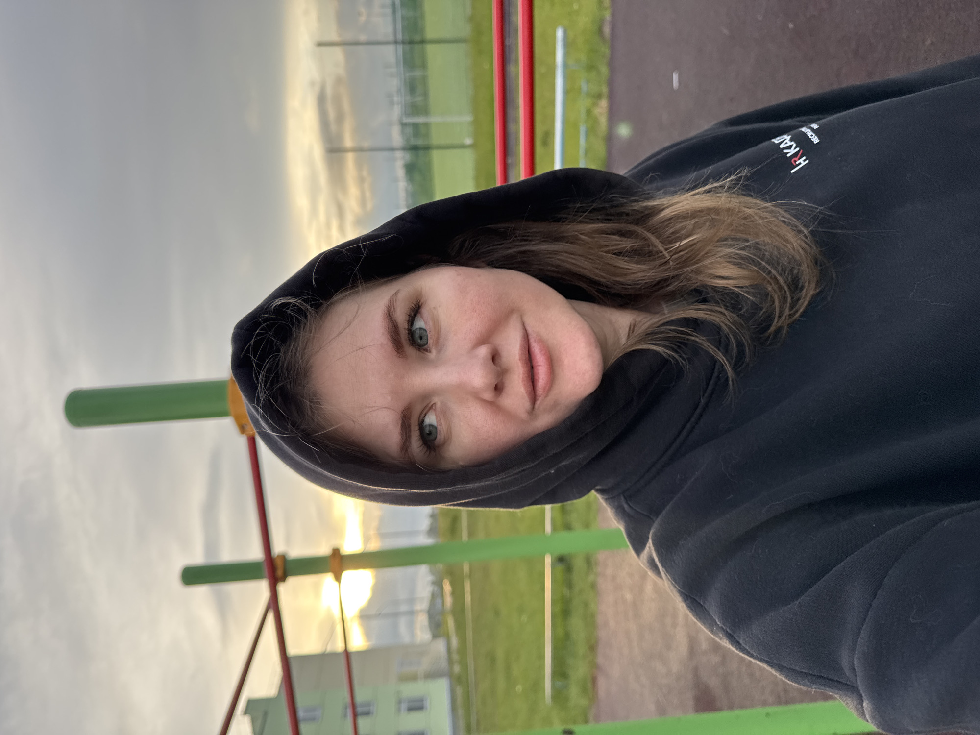 A young adult female, likely in her late 20s or early 30s, is depicted in a medium-close-up outdoor portrait in Горный, Russia. She has fair skin, light brown wavy hair visible under a black hooded sweatshirt with the hood up. Her eyes are blue-green, and she has a small, faint mole on her right cheek (viewer's left). Her lips are slightly parted in a neutral expression, and she is looking to her left, away from the camera. On the left chest of her black hoodie, a small logo composed of white and red text reads "HRKAД" with "RECLAI" or "RECRUI" in smaller white text underneath.
The setting is an outdoor recreation or sports area. In the immediate foreground, a dark reddish-brown textured ground surface is visible. Behind the subject, to the left, are bright green vertical poles and red horizontal bars, part of a playground or exercise structure. Beyond these structures, a multi-story light green building with white window frames is visible. Intense golden light from a low sun or sunset illuminates the sky on the left side, indicating either early morning or late evening. To the right, a large green grass field, resembling a soccer pitch, extends into the distance, featuring white goalposts on the far side. The sky above is largely overcast with grey clouds, interspersed with areas of lighter, golden-hued cloud cover on the horizon.