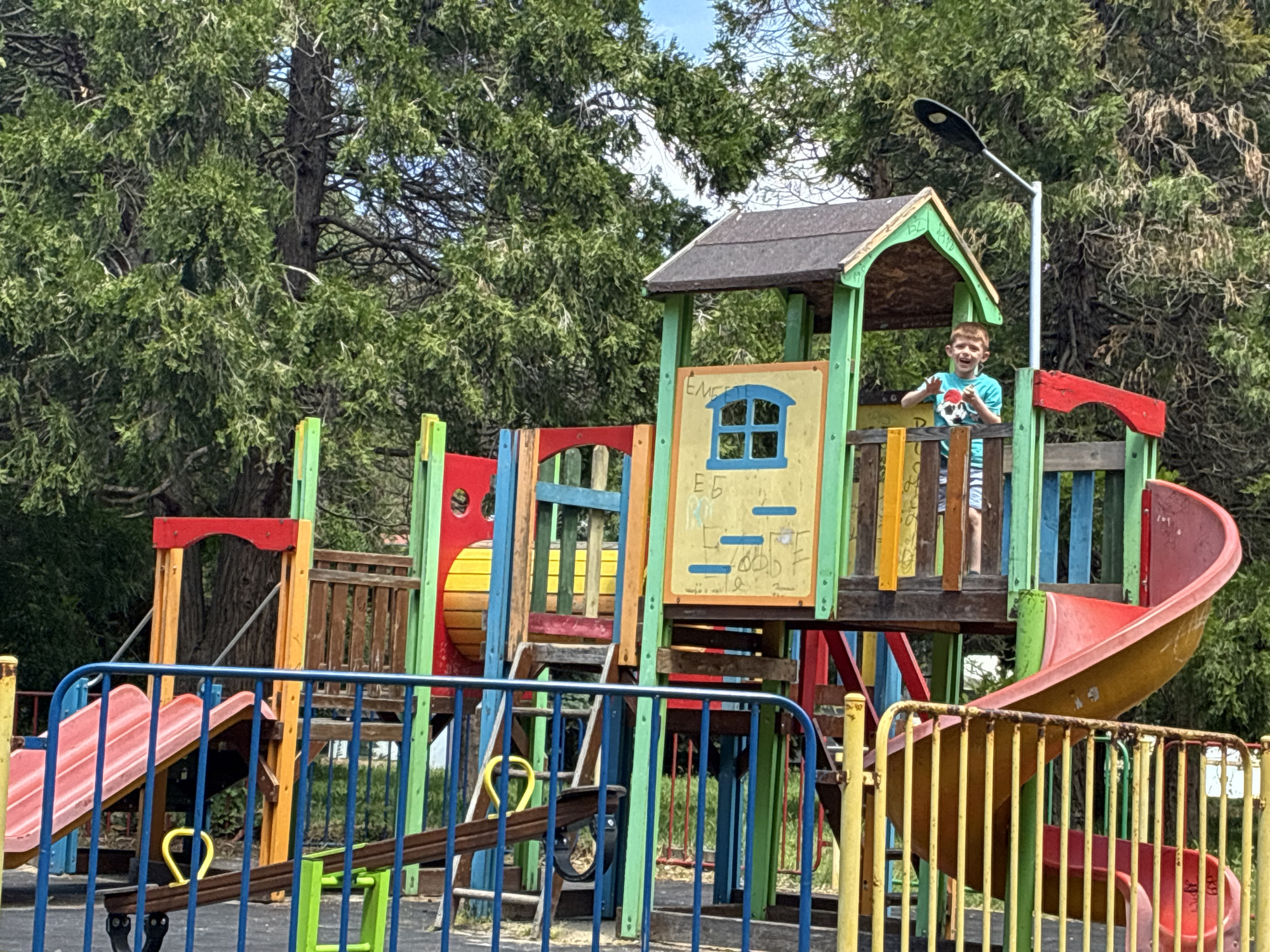 A playground in Varna, Bulgaria, features a colorful, multi-tiered play structure. A young boy with short red hair, wearing a blue t-shirt with a skull design, stands atop the structure, waving. The structure includes slides, ladders, and fenced platforms. A light post stands adjacent to the structure. Blue, yellow, and green metal fencing surrounds the playground. Large trees are in the background.