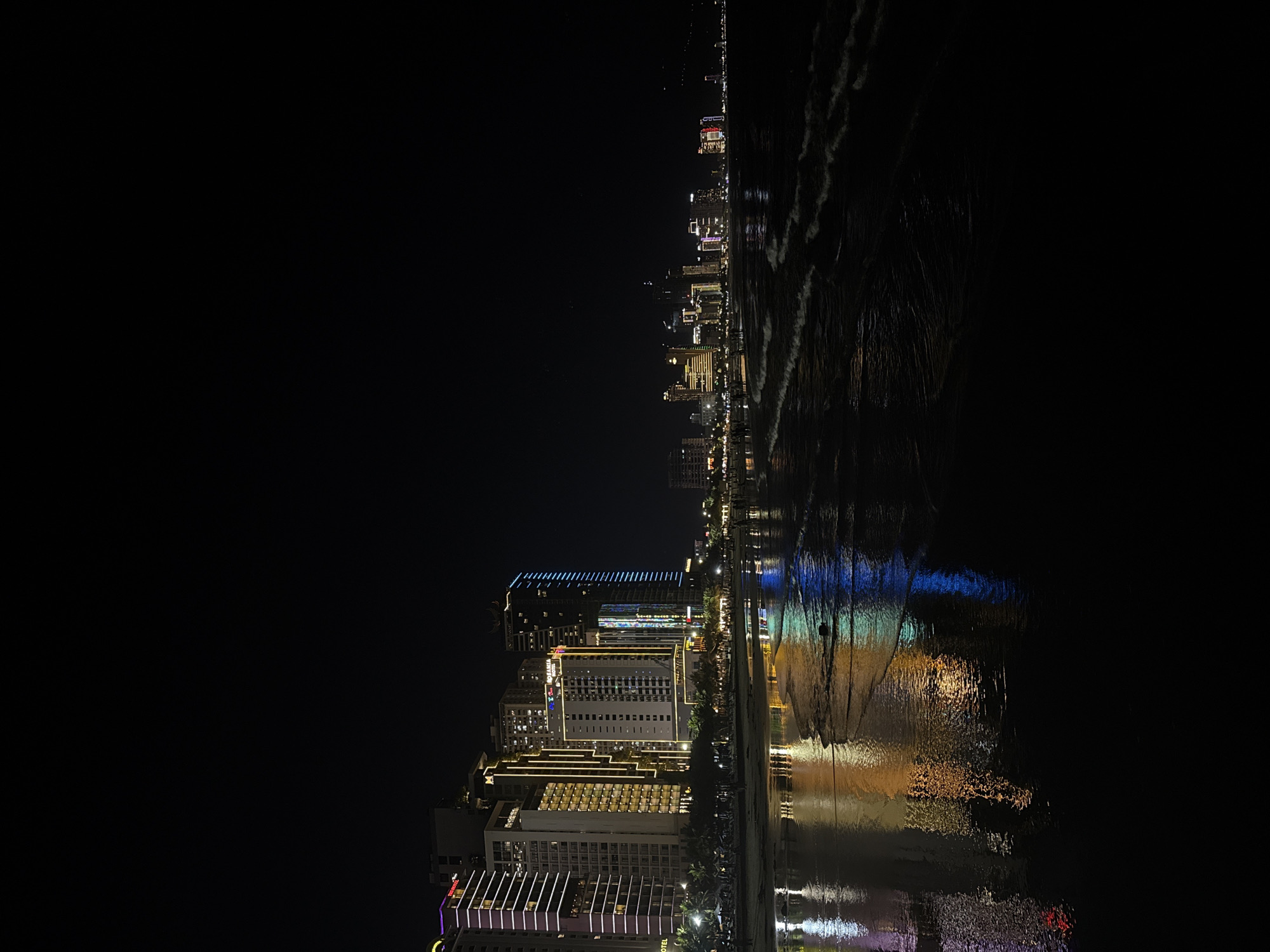 The image captures a nighttime scene of the beachfront in Da Nang, Vietnam, facing a row of illuminated high-rise buildings. The dark sky is punctuated by the bright, colorful lights of the hotels and businesses, which are reflected in the wet sand and shallow water along the shoreline. Several waves gently break on the beach, their crests catching the ambient light. People are visible as small silhouettes walking along the beach in the mid-ground. The buildings are a mix of modern architecture, featuring various lighting designs from subtle white and yellow to vibrant blue and pink stripes. The overall impression is of a lively, urban coastal city at night.