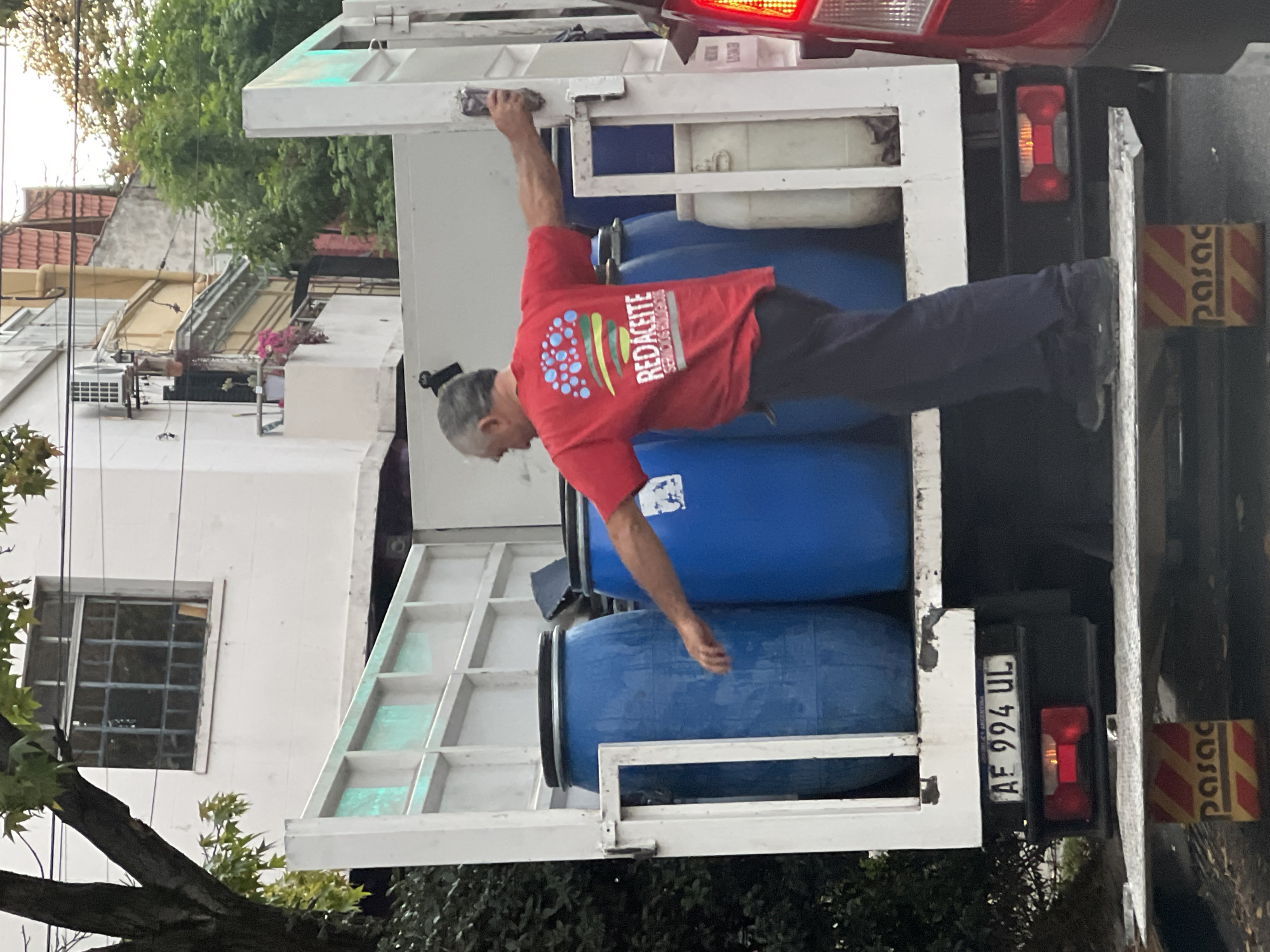 A man in a red t-shirt with the word "REDACEITE" stands in the back of a truck filled with large blue barrels. He is holding onto the top of the truck with his left hand and reaching towards the barrels with his right hand. The truck's license plate reads "AE 994 UL," indicating it is in Argentina. The surrounding environment suggests a residential area in Buenos Aires, Argentina.