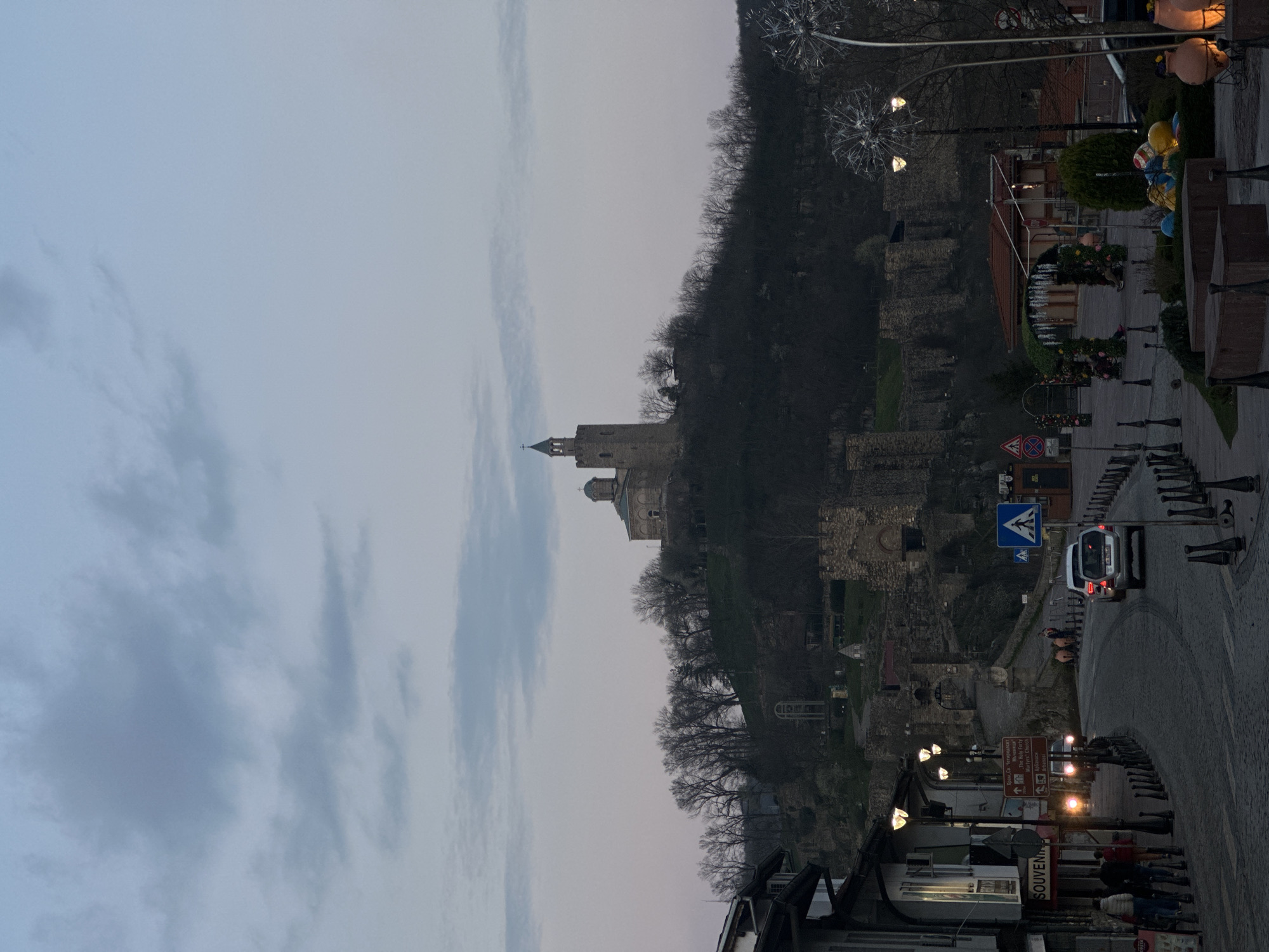 The image depicts the Tsarevets Fortress in Veliko Tarnovo, Bulgaria, at dusk. The fortress, featuring stone walls and a tower with a cross, sits atop a hill. The sky is overcast with light grey clouds. Below the fortress, there's a street with visible pedestrian and vehicular traffic. Streetlights illuminate the road and buildings. A white van is driving along the road, and several people are walking on the sidewalks. "Souvenirs" is written on the building. A tourist board indicates "The Holy Forty Martyrs Church". The setting is a blend of historical architecture and contemporary urban life.