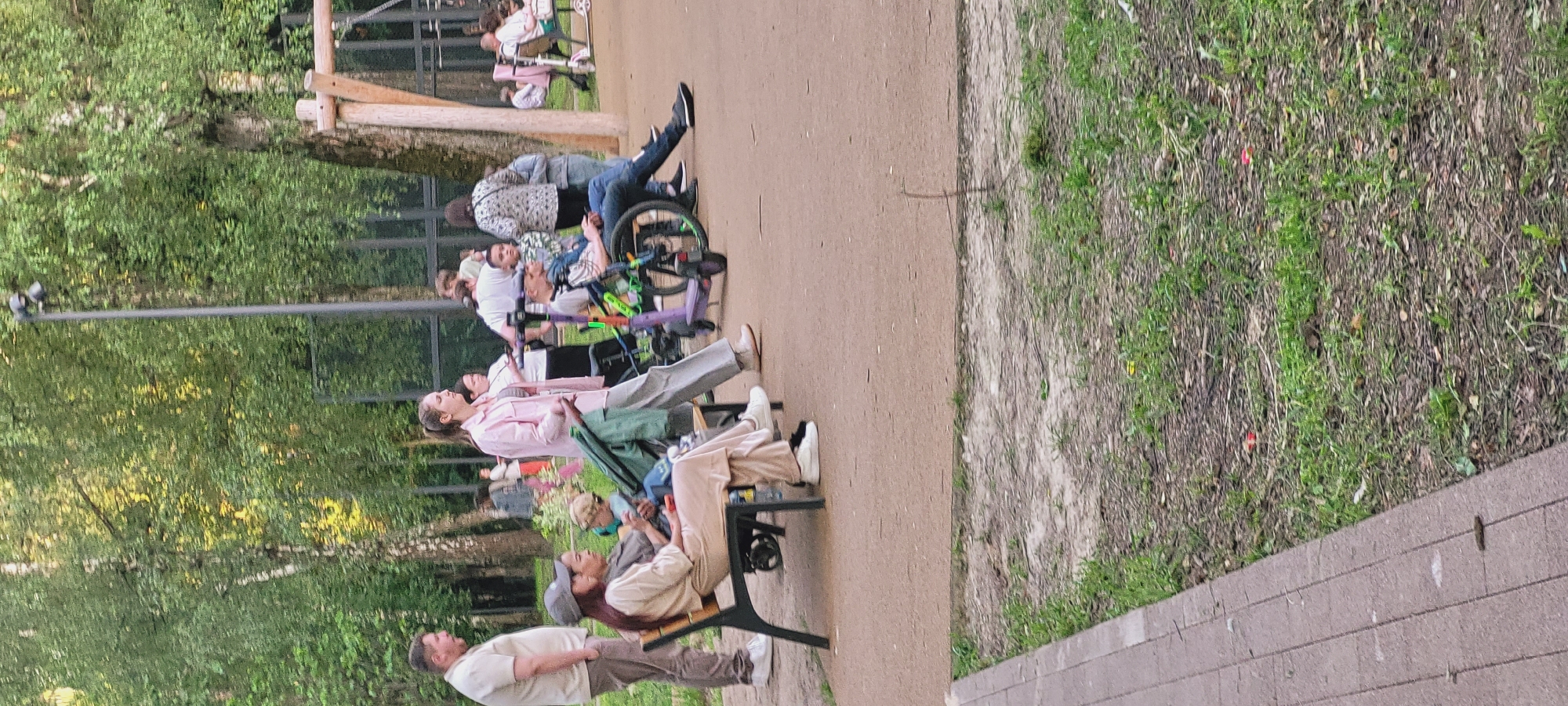 The image captures an outdoor scene in a public park or recreational area within the city of Unknown, Unknown. The ground consists of compacted light brown earth with scattered patches of green grass and small debris. A grey concrete paver path is visible along the bottom left edge of the frame.
In the foreground to mid-ground, a group of individuals is present. On the left, a light-skinned male wearing a light tan short-sleeved shirt and matching trousers stands with his hands in his pockets, looking right. Adjacent to him, a light-skinned woman with long dark hair, partially covered by a grey baseball cap, is seated on a black metal bench. She wears a light beige long-sleeved top and matching wide-leg trousers, with her legs crossed and a small red object in her hand. A second person, partially obscured behind her on the bench, wears a grey top. A green and black child's stroller is positioned near the bench, containing a light-haired child looking right.
To the right of the bench, a tall light-skinned woman with a ponytail stands, wearing a light pink long-sleeved shirt over a striped top and light grey wide-leg trousers. She is looking left. Further to the right, a light-skinned male, likely a young adult, is seated in a black and green manual wheelchair. He wears a grey t-shirt and blue jeans, with his left leg extended forward. A purple and green kick scooter rests near the wheelchair.
In the mid-ground, several other individuals are visible. These include a light-skinned woman in a white t-shirt and dark pants, a child in a patterned shirt, and another woman in a patterned top. The background features numerous deciduous trees with dense green foliage, including a prominent birch tree trunk. A tall dark utility pole with two light fixtures is situated in the mid-background. Black mesh fencing runs horizontally across the mid-ground, and a partial wooden structure, possibly playground equipment, is visible on the far right. The lighting is soft, suggesting an overcast day or late afternoon.