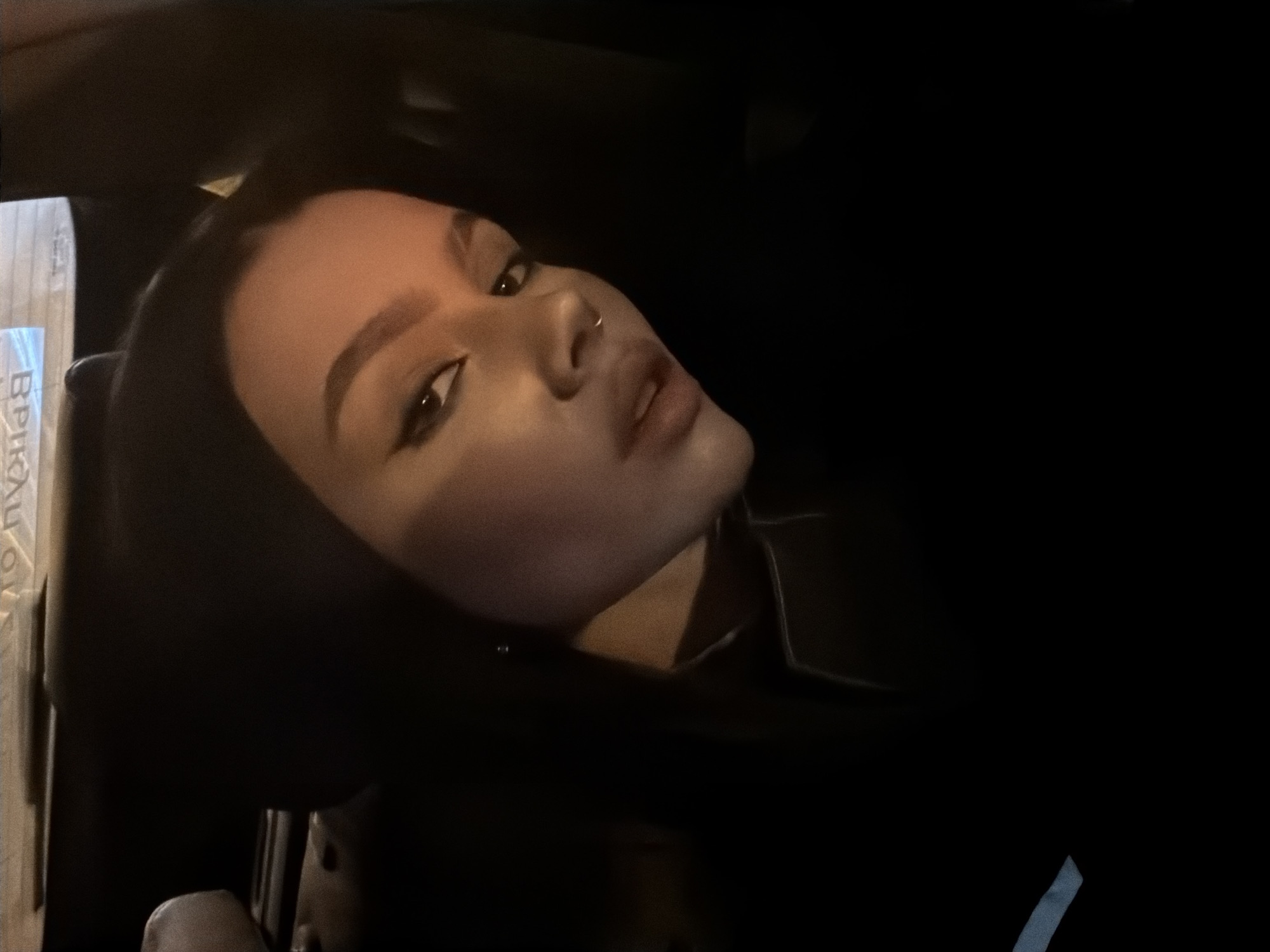 A close-up, dimly lit photograph shows a person with fair skin, dark hair, and dark eyeliner, likely female, with a nose ring. The person is positioned at an angle, looking up and to the left. The surrounding environment is dark, possibly inside a car or other enclosed space. There are indistinguishable objects out of focus on the left side of the frame, and a small strip of white or light-colored material is visible in the lower right corner. No details are present in the image to suggest the city of Moscow, Russia.