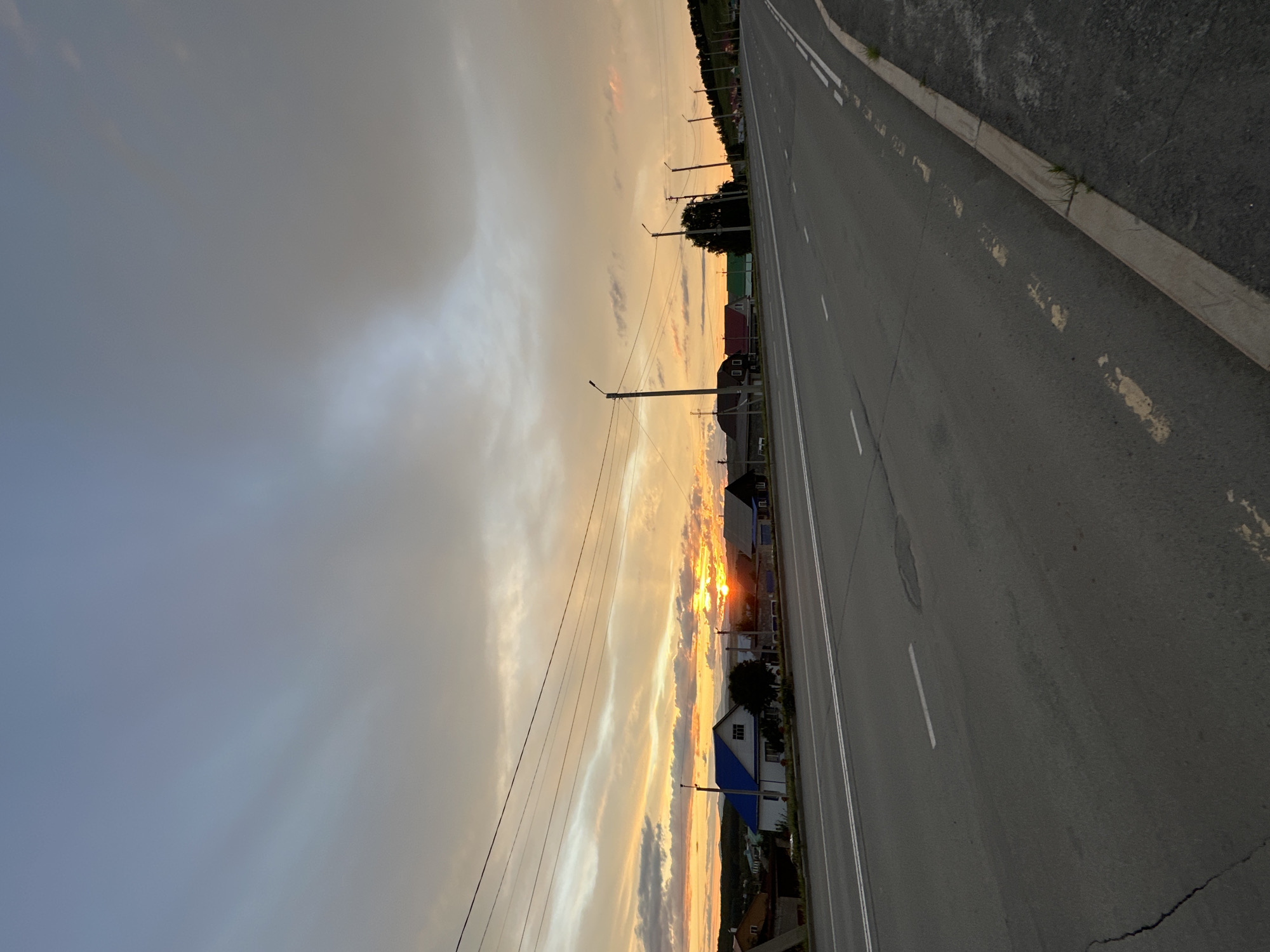This is a photograph taken at dusk of a straight, paved road with double white lines. The road curves to the right in the distance. On the left side of the road, a row of houses with pitched roofs can be seen, some with blue roofs. Streetlights are visible along the road. The sky is filled with clouds, with a bright orange and yellow sunset visible through them, casting a warm glow. Power lines are strung across the upper part of the image. The ground on the right side of the road is rough and uneven with some sparse vegetation.