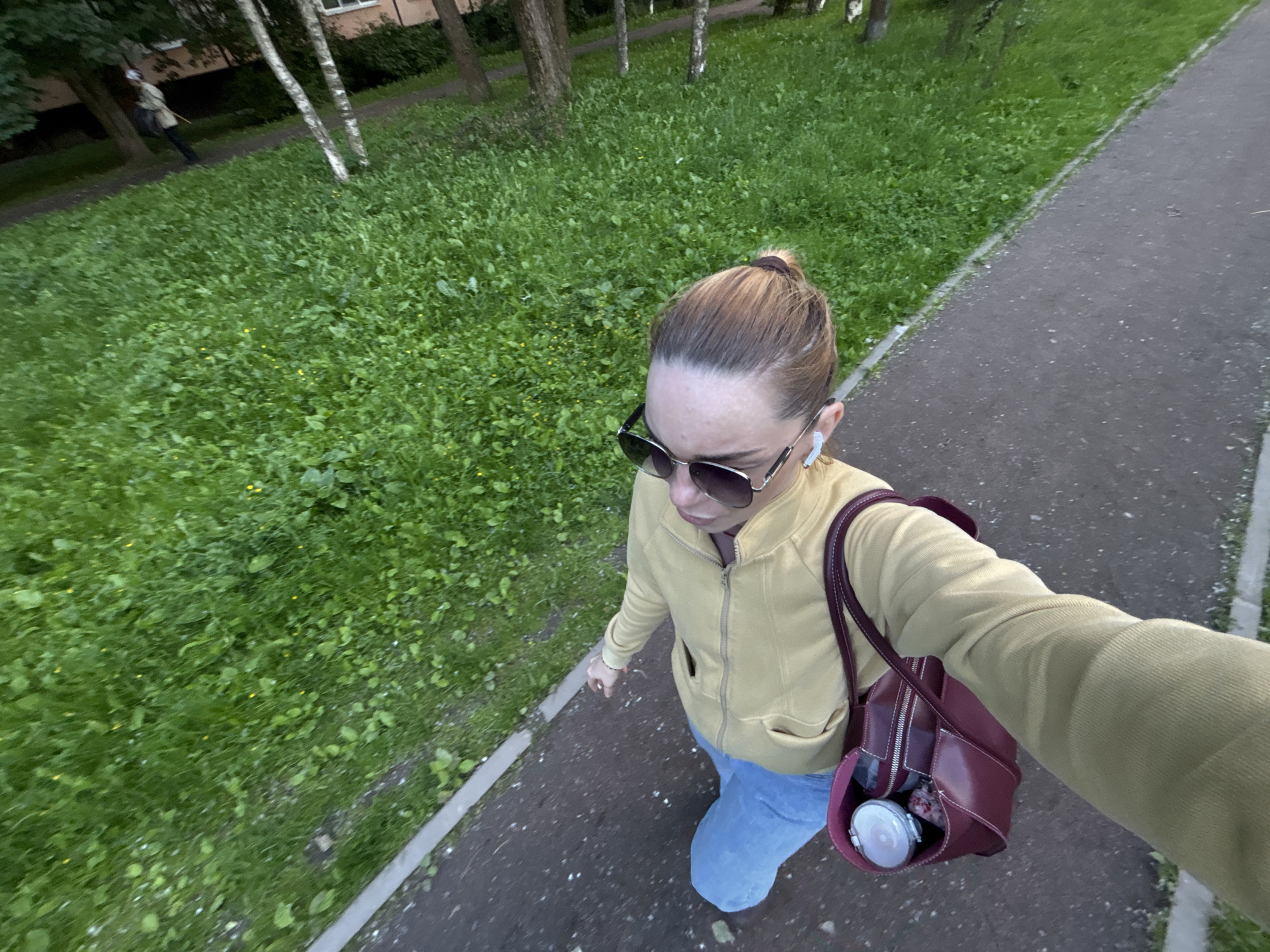 A young woman is walking on a paved path in Sinyavino, Russia, with a patch of lush green grass to her left. She is wearing a yellow zip-up hoodie, light blue jeans, and sunglasses. A burgundy handbag hangs from her right shoulder, containing a white object that appears to be headphones. She has her hair pulled back in a bun. The path is bordered by the grassy area and appears to be a walkway in a residential or park setting. In the background, trees and a building are visible. Another person, dressed in lighter clothing and carrying a stick, is walking away from the camera in the distance on the grassy area.