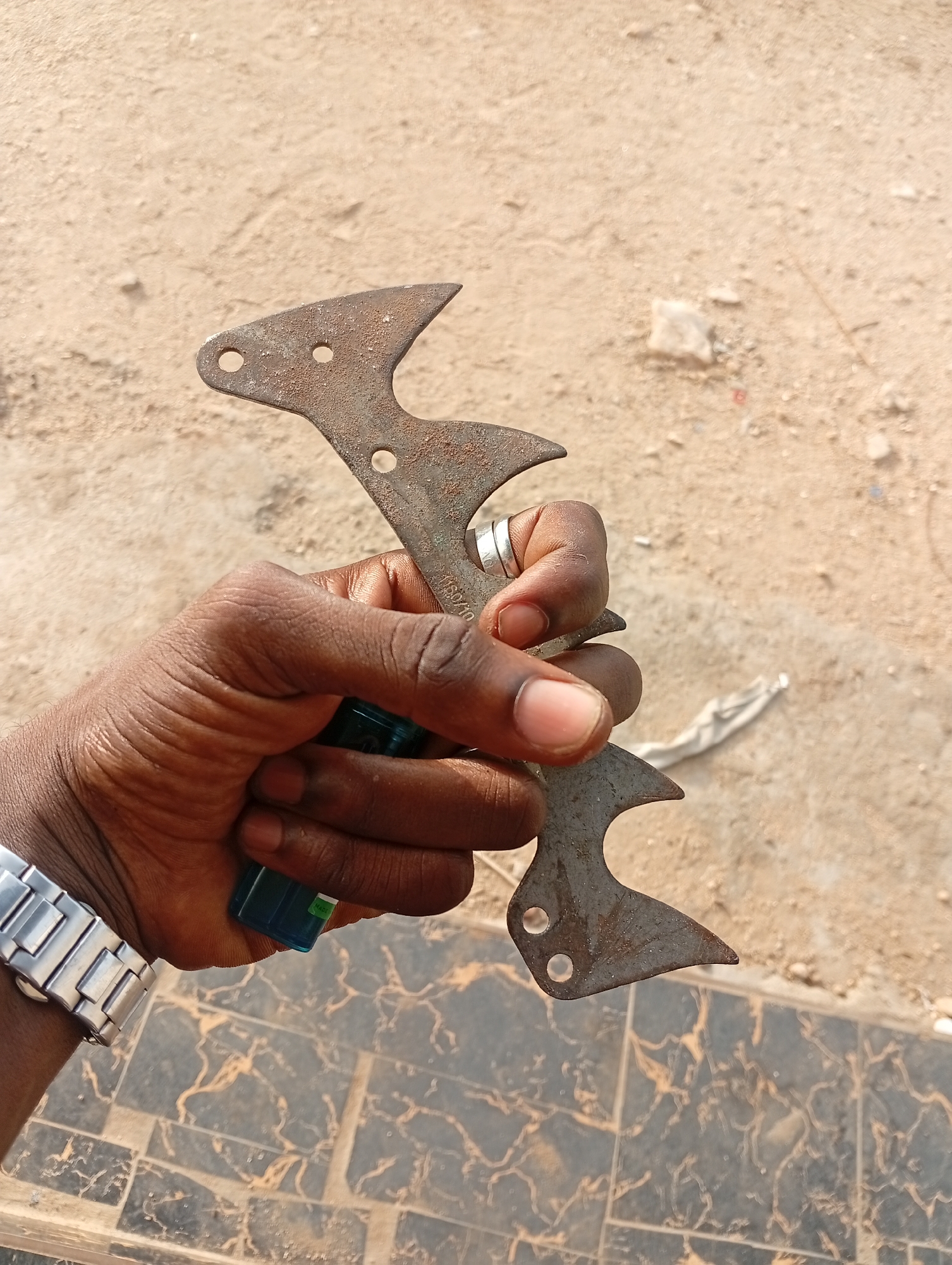 An arm, presumably male, wearing a silver watch is holding a rusty metal tool. The tool is flat with three holes at each end. The hand holding the tool wears a silver ring. The environment is outdoor. A beige, sandy area appears in the background. A tiled surface with a dark marble pattern is beneath the arm. The location is assumed to be Adankolo, Nigeria based on the user prompt.