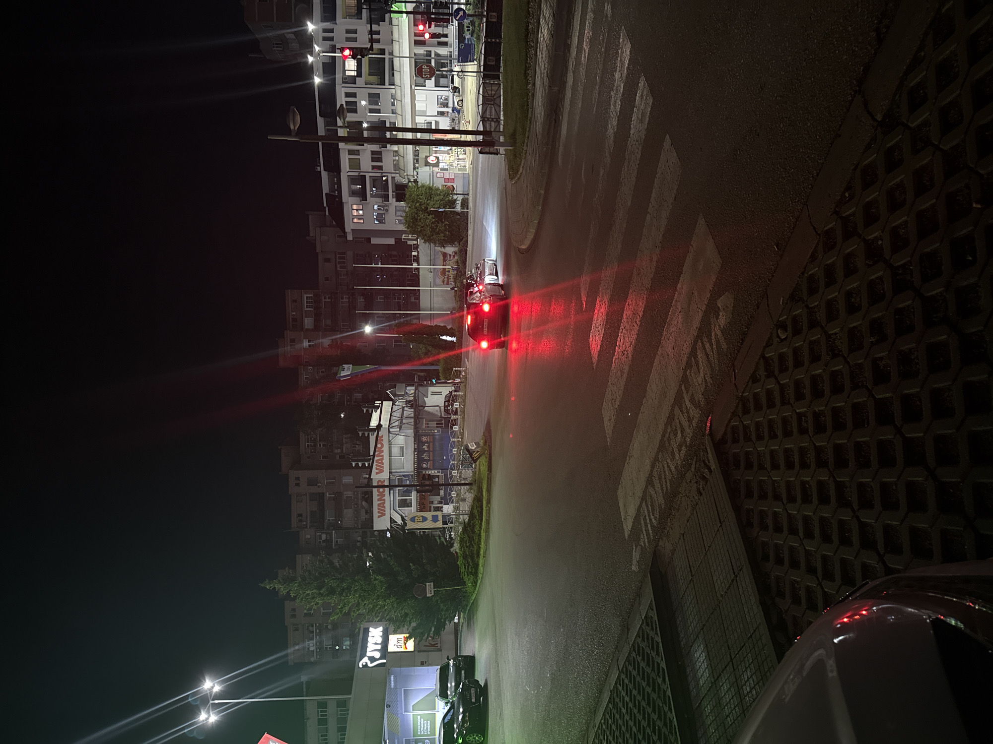 The image depicts a nighttime street scene in Veliko Tarnovo, Bulgaria. A car with illuminated red tail lights is positioned in the middle of the street, facing away from the viewer, near a zebra crossing. The street is wet, reflecting light. Buildings line the left side of the street, some with lighted windows. A JYSK store is visible on the left, with signage lit up. Streetlights illuminate the scene. A car is parked near the curb on the left. The pavement is visible on the right. Pedestrian crossings are visible. A stop sign is illuminated in the distance. Traffic lights are also visible. The image is taken at an oblique angle. The street appears to curve.
