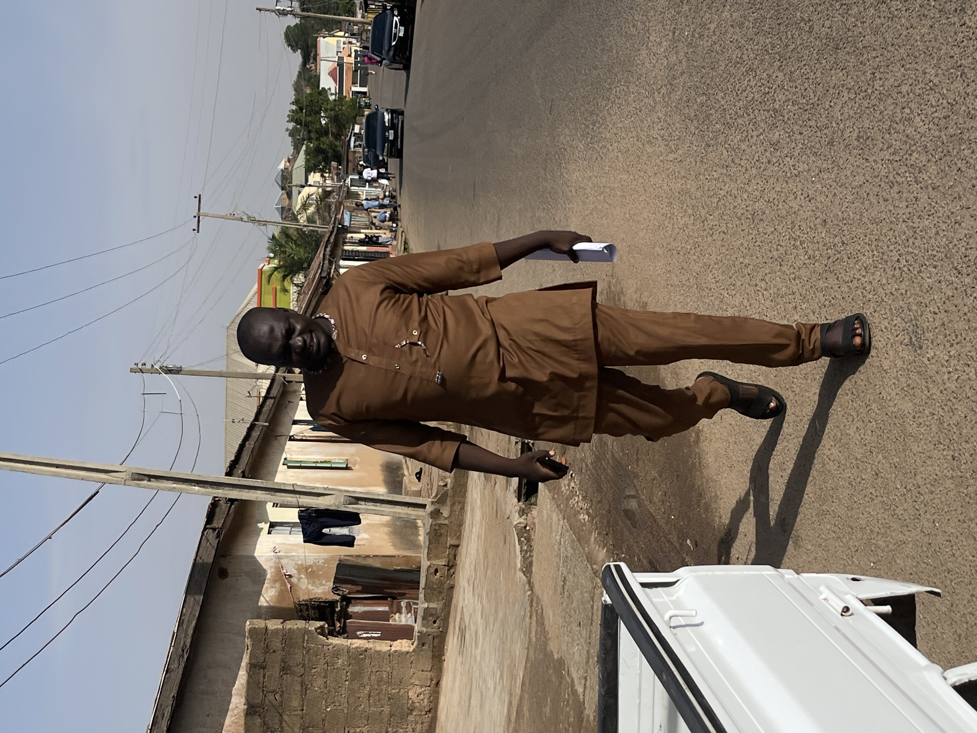 A man stands on a street in Jos, Nigeria. He wears a brown, long-sleeved outfit with matching pants and sandals. He holds a white, rolled-up paper in his right hand. The street behind him shows cars and pedestrians. Buildings are visible on the left, with clothing hanging outside one. Power lines stretch across the sky. A white vehicle is partially visible in the foreground.