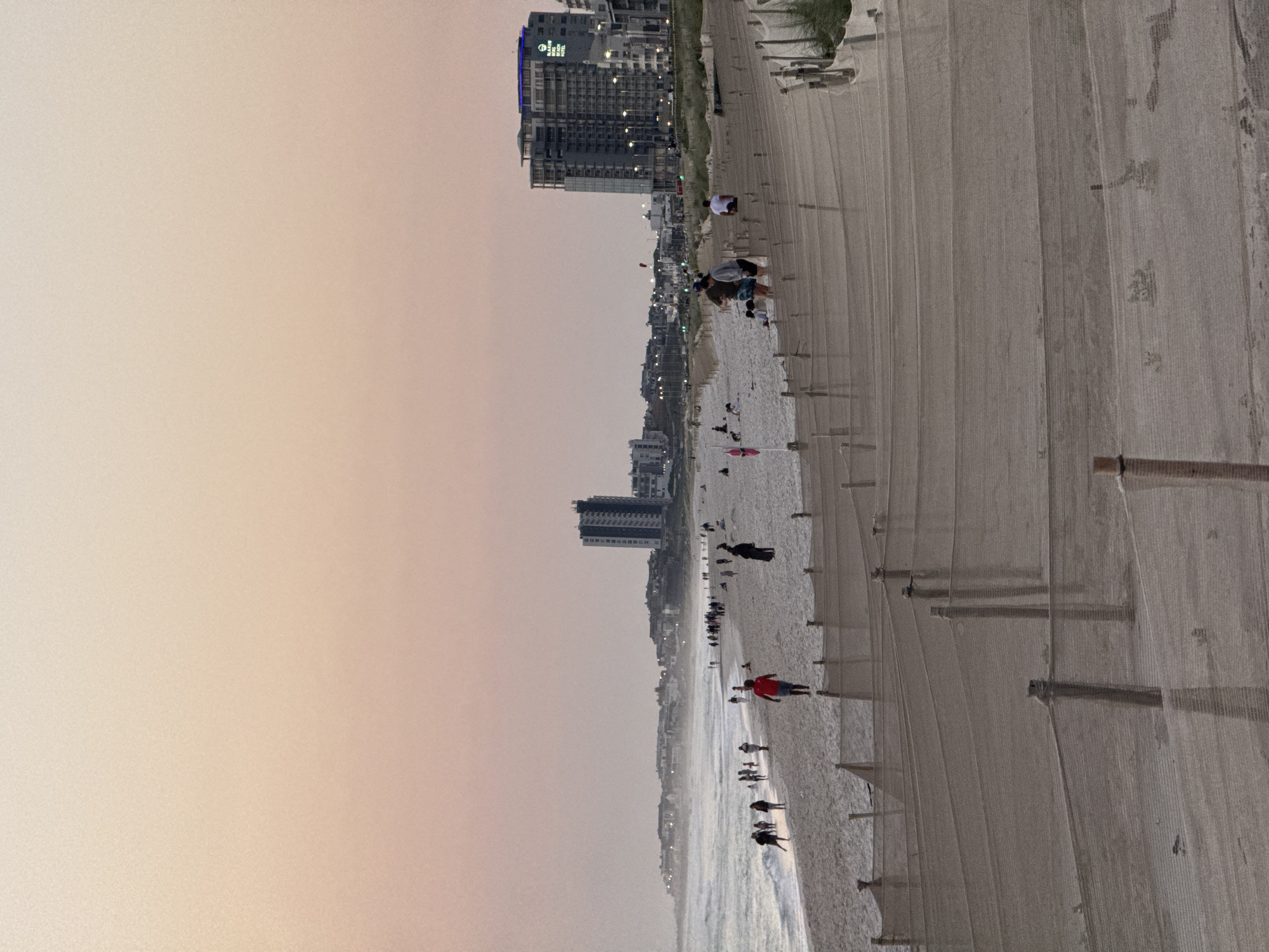 The image depicts a wide, sandy beach in Cape Town, South Africa, during what appears to be either sunrise or sunset, given the soft, pastel hues of the sky. Numerous individuals are present on the beach, engaged in various activities such as walking, standing, and seemingly interacting with each other. In the foreground, a series of low fences, constructed with netting and wooden posts, are visible, possibly serving as a demarcation or protection barrier. Beyond the beach, a coastline stretches into the distance, featuring a cluster of buildings, including a prominent tall hotel with a sign that reads "BLAUWBERG BEACH HOTEL." The ocean is visible on the left side of the image, with gentle waves rolling towards the shore. The overall atmosphere is serene, with a sense of public recreation and coastal living.