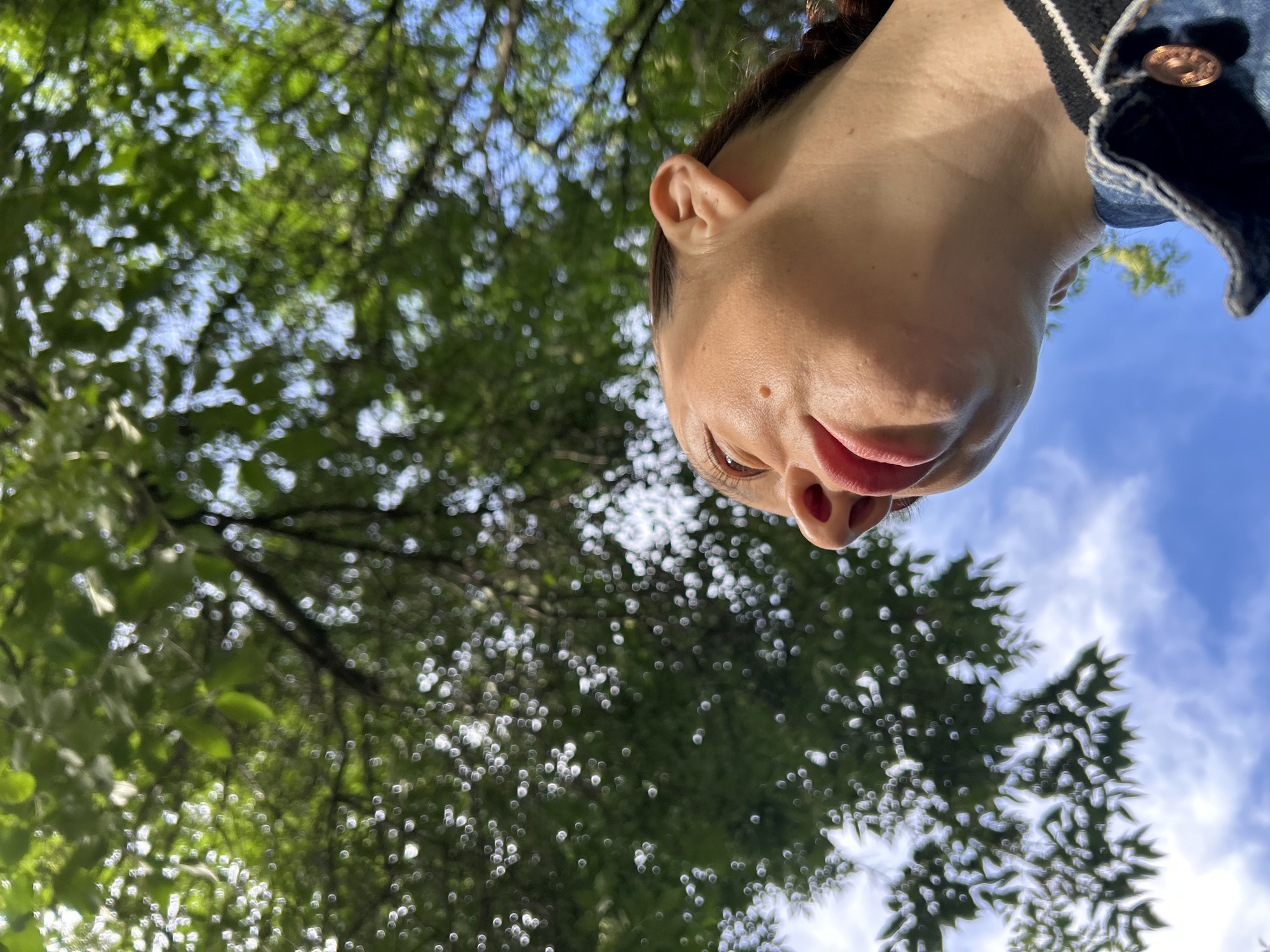 The image shows a close-up, low-angle shot of a person's face, specifically their nose and mouth, looking upwards. The background is dominated by the dappled green foliage of trees and a bright blue sky with scattered white clouds. The lighting suggests it is daytime. The person is wearing a denim jacket. There is no information in the image to indicate the location in the city of Chaikovsky, Russia.