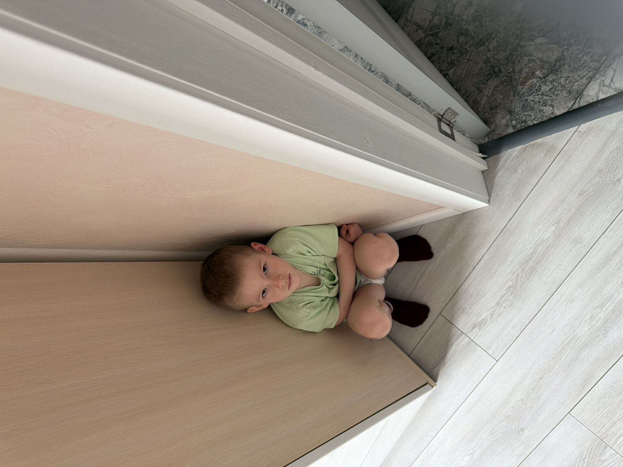 A child sits between a wall and a cabinet. The child is wearing a green shirt and dark red socks. The walls are a light peach color, and the cabinet is a light wood color. The floor is tiled in white and gray. The location is inferred as Rossiyskiy, Russia, based on the user query.