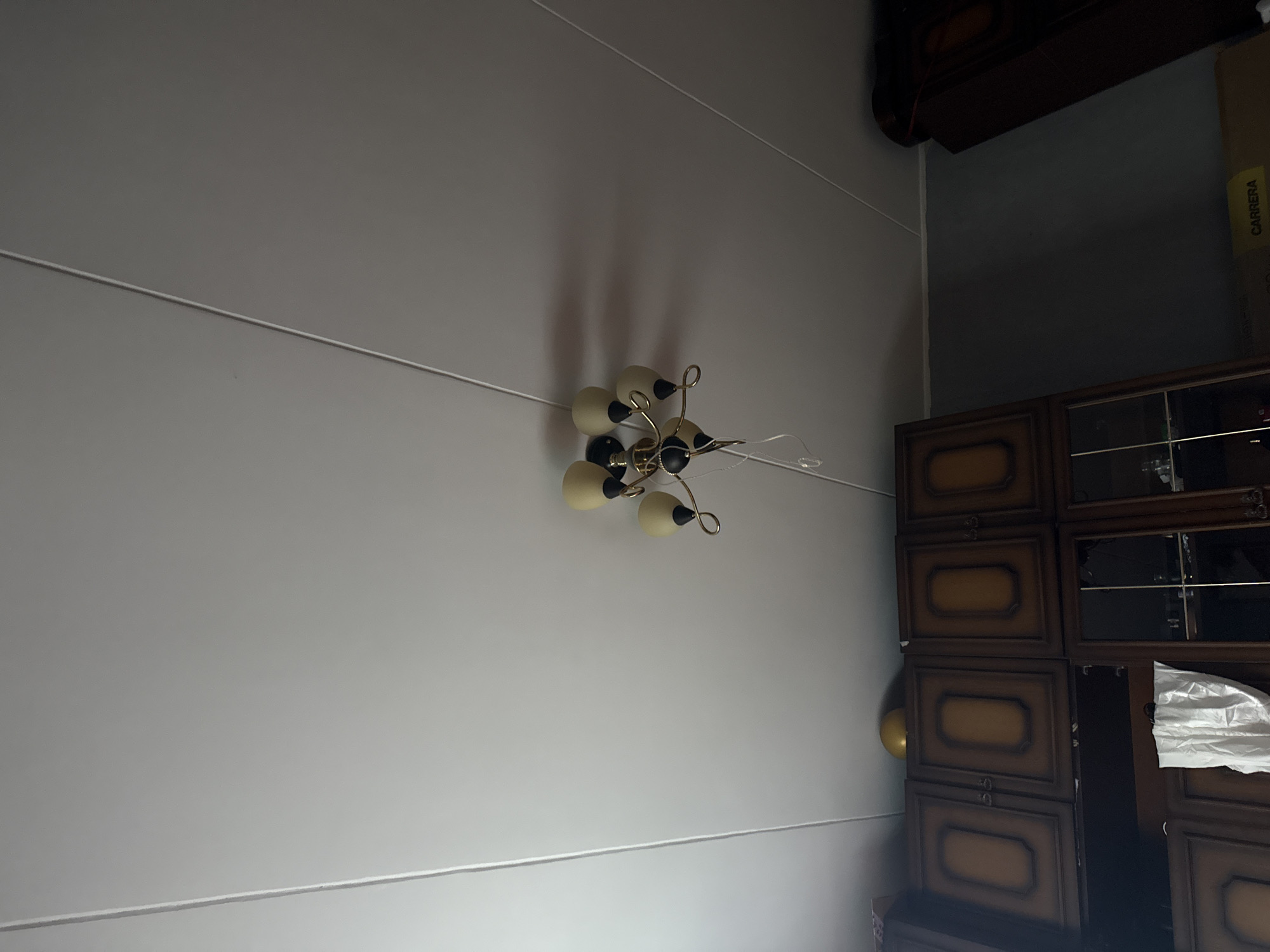 This is an interior shot of a room, likely a residential dwelling, taken from a low angle looking up at the ceiling. The ceiling is white and appears to be made of paneling, with visible seams running diagonally across the frame. A vintage-style chandelier with multiple light fixtures hangs from the ceiling. The chandelier has a gold-colored frame and cream-colored lampshades. Below the ceiling, a portion of a dark wooden cabinet or bookshelf is visible. The cabinet has ornate detailing and several doors. On top of the cabinet, a single yellow balloon is placed. To the right of the cabinet, a dark wall is visible. The overall impression is of a room with somewhat dated furnishings. There is no information in the image to suggest this location is in Moscow, Russia, or any other specific city.