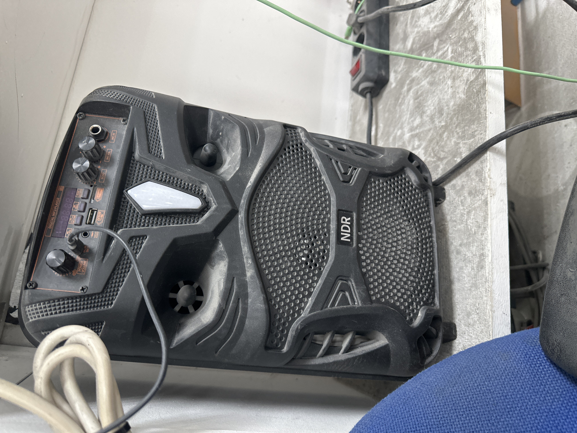 The image shows a black portable speaker with the brand name "NDR" visible on the front. The speaker has a mesh grille, control knobs, and various ports on its top panel. It is positioned on a dusty, textured surface, likely a table or shelf, in an indoor environment. A blue chair is partially visible in the foreground. Several cables, including a thick white cable and a thin green cable, are connected to or near the speaker and extend out of the frame. The background is a plain, light-colored wall. The scene is static, with no discernible actions or interactions taking place. The overall impression is one of a utilitarian setting, possibly a workshop or storage area. The location is not specified as Mainz, Germany.